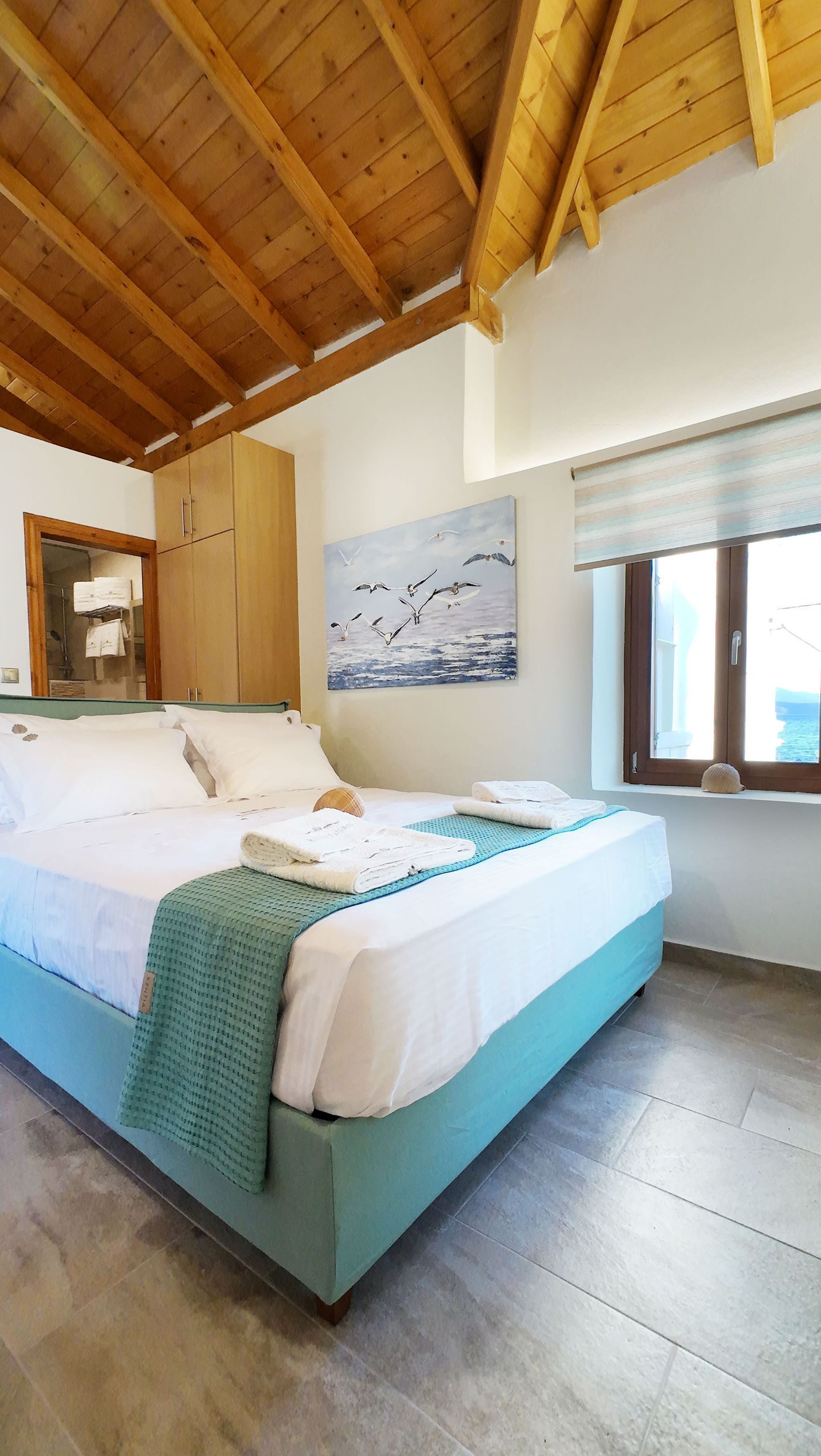 Een slaapkamer met een bed, houten balken in het plafond, een schilderij van vogels, een raam met uitzicht en een betegelde vloer.