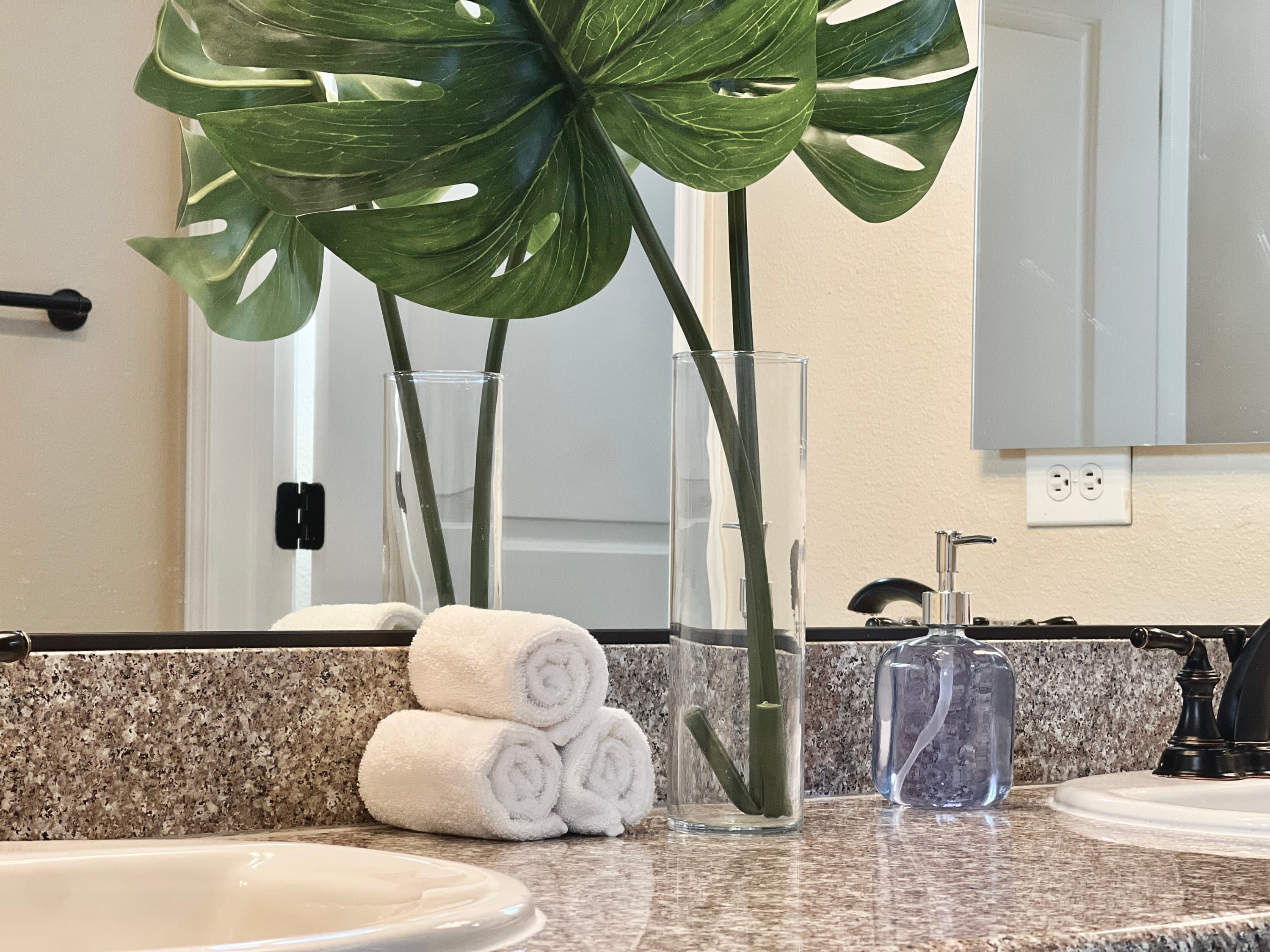 Une salle de bain avec un comptoir en granit, deux vases transparents contenant des plantes vertes, un lavabo et un miroir.