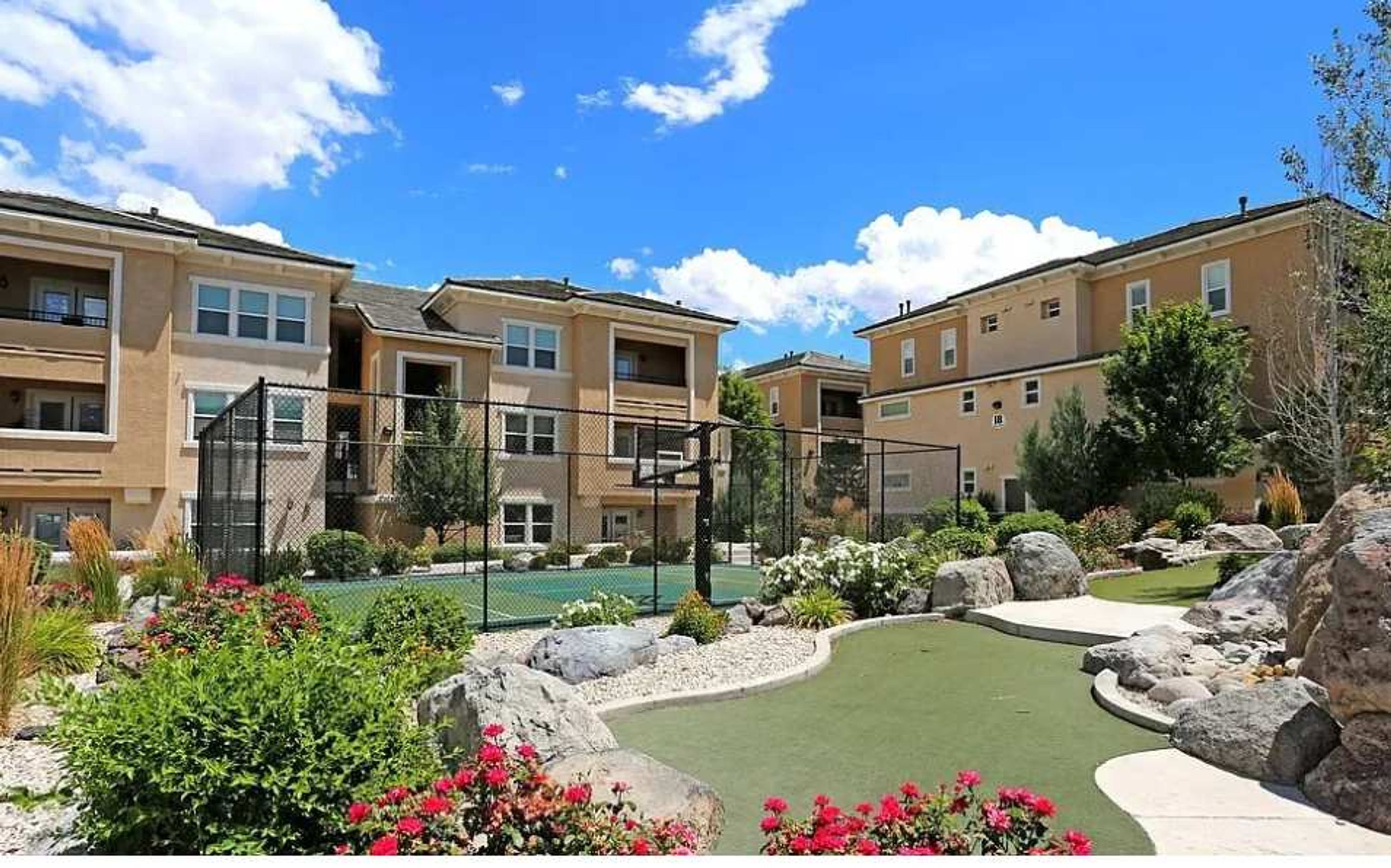 A residential complex with multiple apartment buildings, a fenced tennis court, and landscaped gardens with rocks and shrubs.