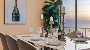A dining table set with plates, glasses, and candles, with a framed Versace bottle on the wall and a large plant in the background. - Sunset View Luxury 4 Bedrooms With Sauna (Mount Martha)