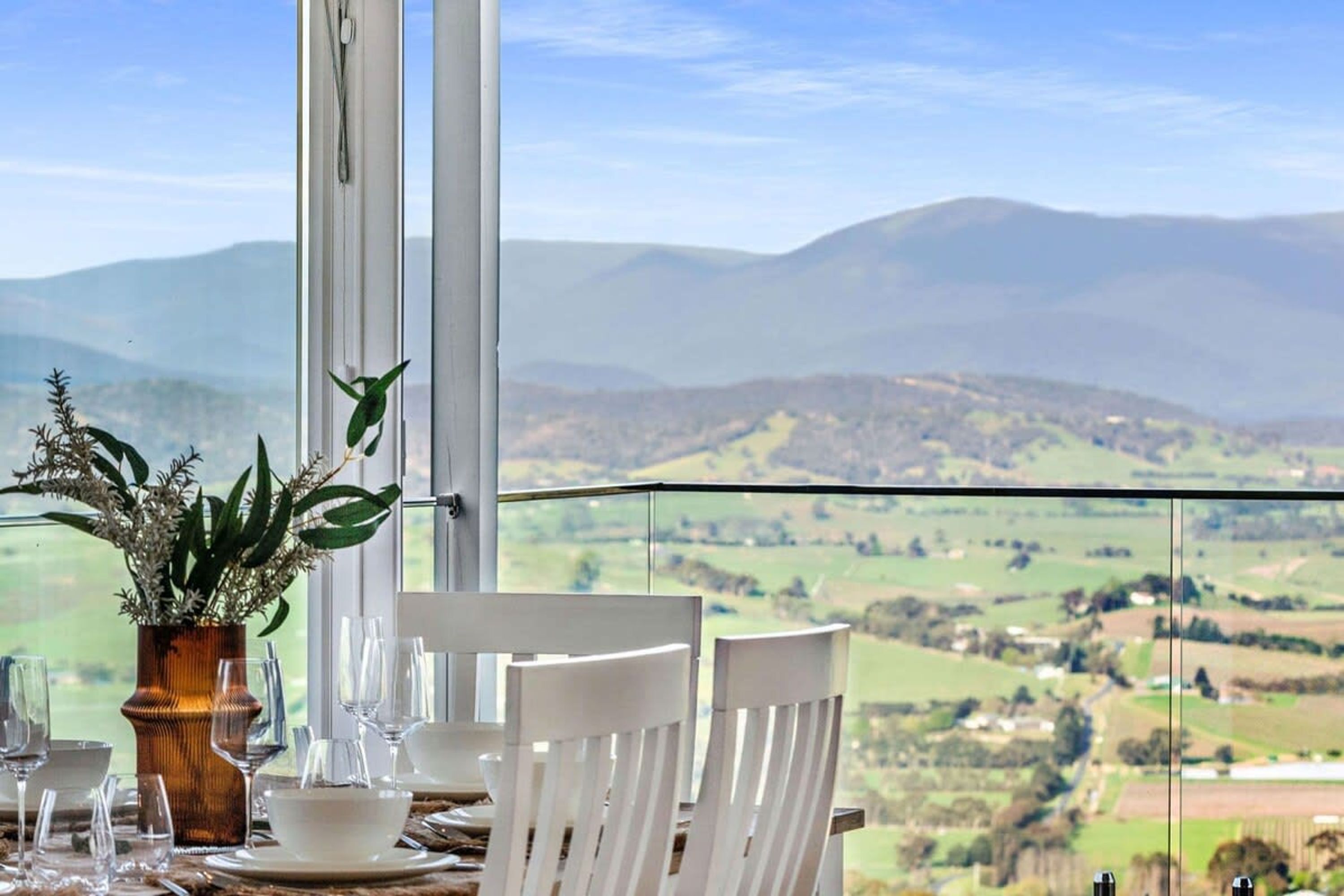 A dining table with white chairs and a vase of flowers, set against a scenic mountain view.