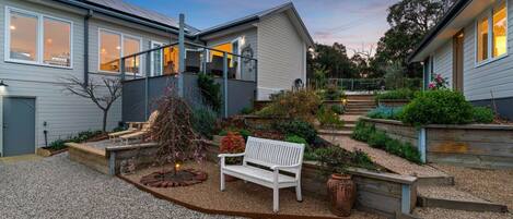 A residential property with a gravel driveway, a white bench, and a small garden area with various plants and a tree.