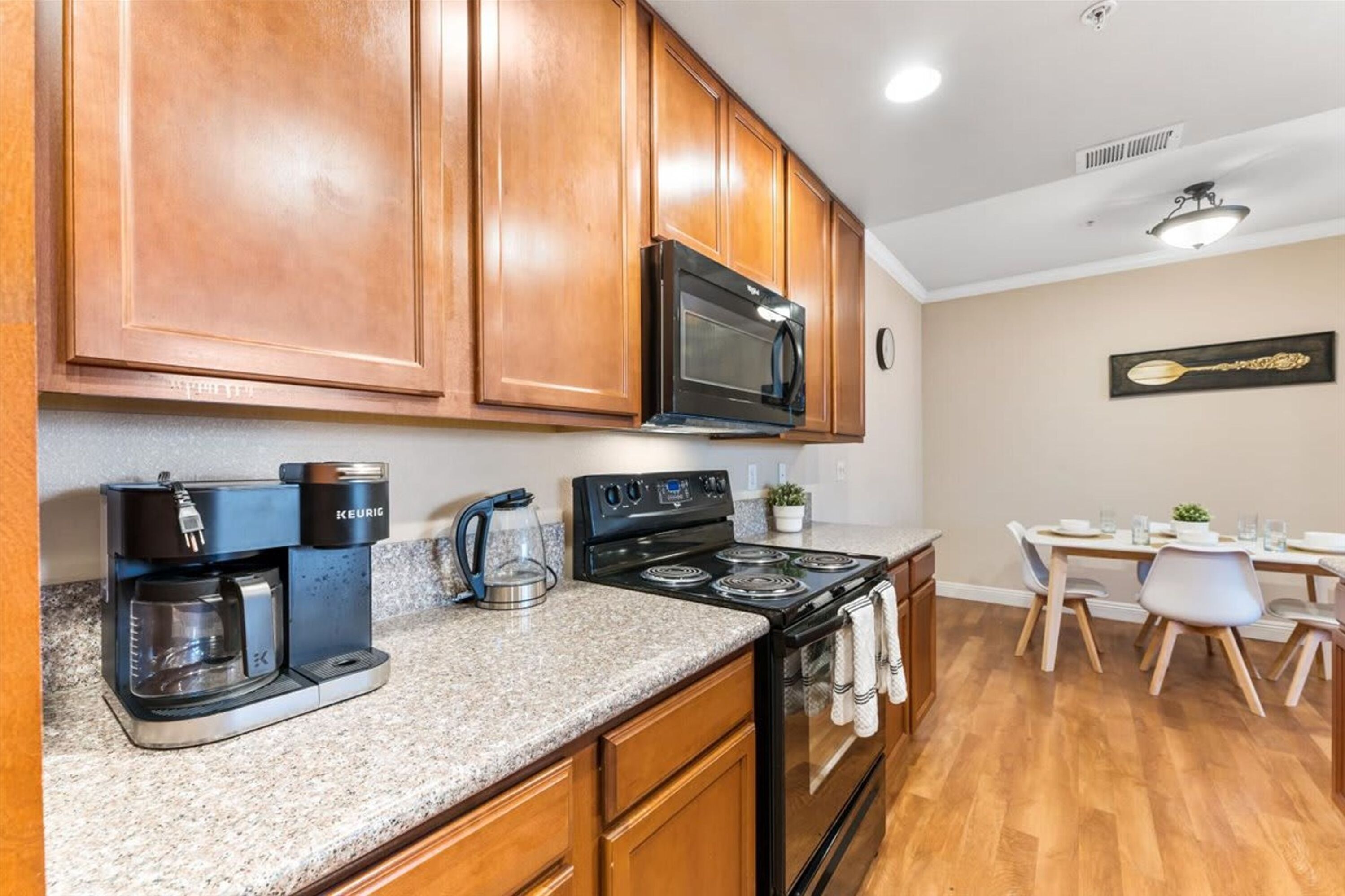 A kitchen with wooden cabinets, a black microwave, a coffee maker, and a stove. There is a dining table with chairs and a plant on the counter.