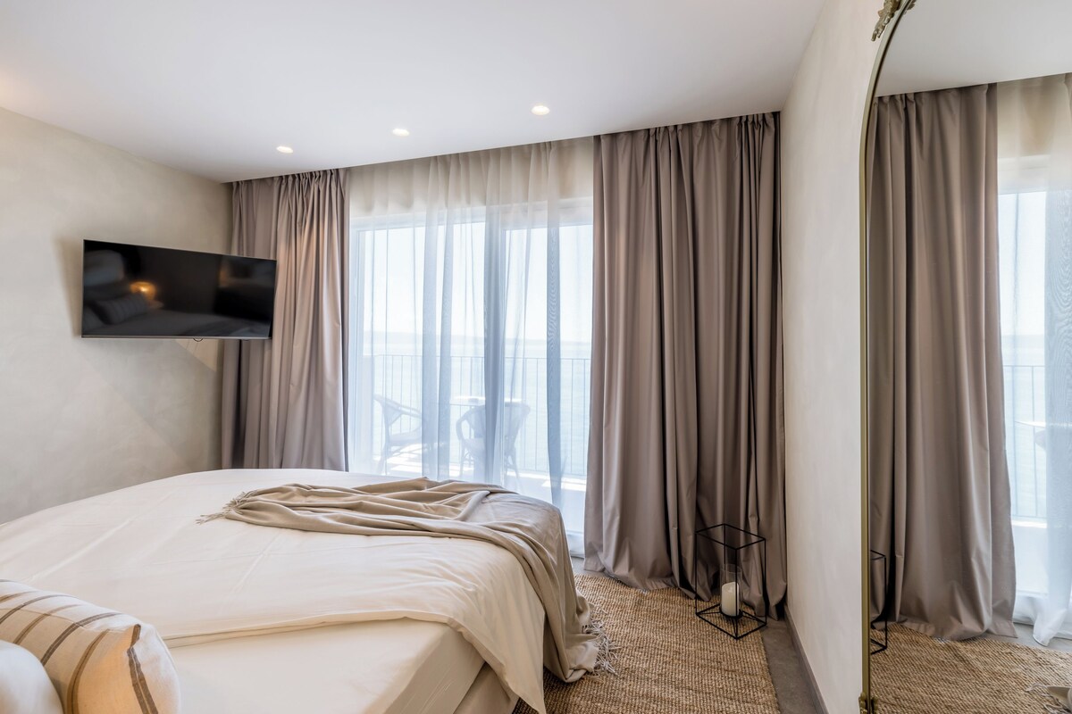 Habitación de hotel moderna con una cama grande, un televisor de pantalla plana en la pared y vistas al exterior a través de cortinas translúcidas.