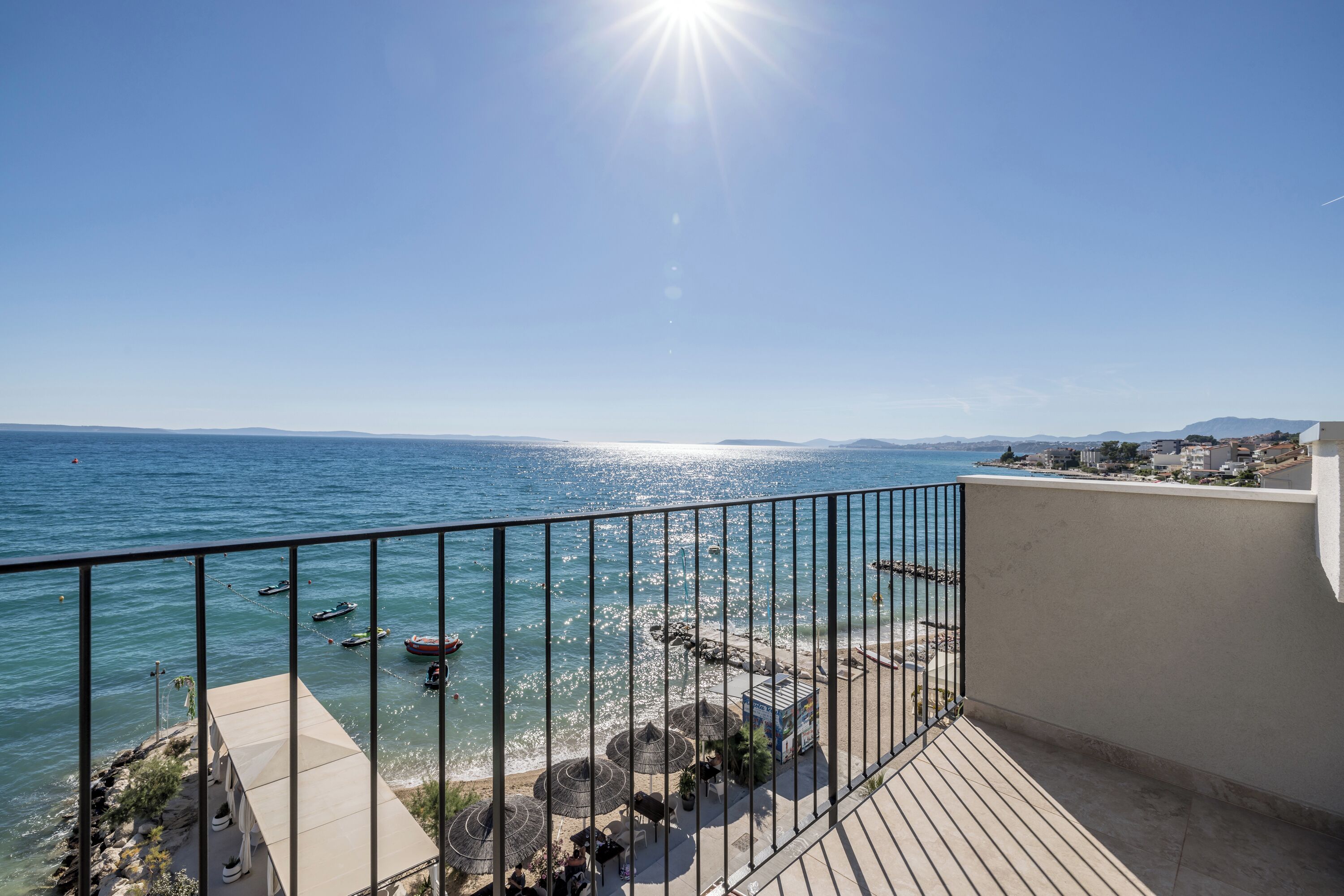 A balcony with a view of the sea, boats, and a beach area with umbrellas and tables.
