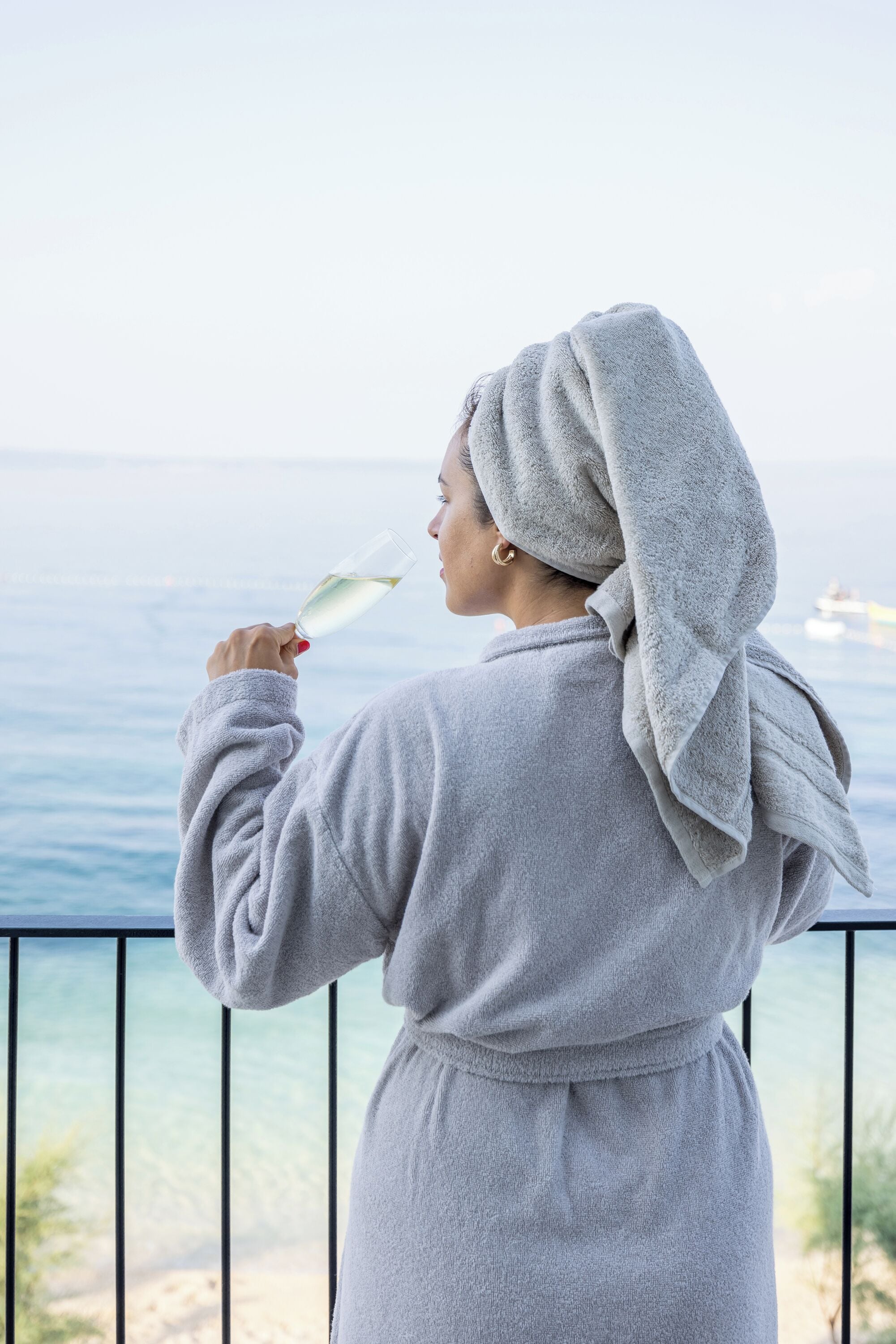 Una persona con una bata gris sosteniendo una copa de vino blanco, de pie en un balcón con vista al mar.