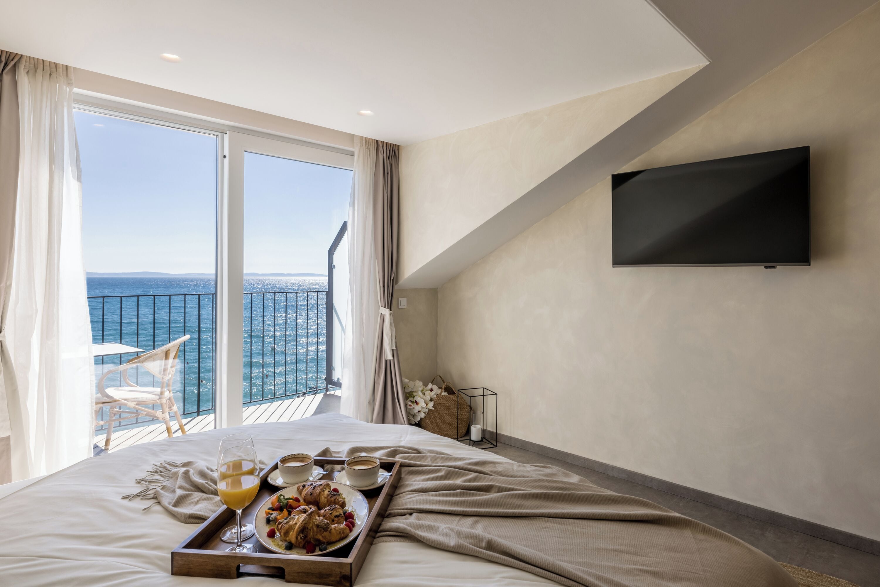 A bedroom with a large bed, a TV mounted on the wall, and a balcony with a view of the sea.