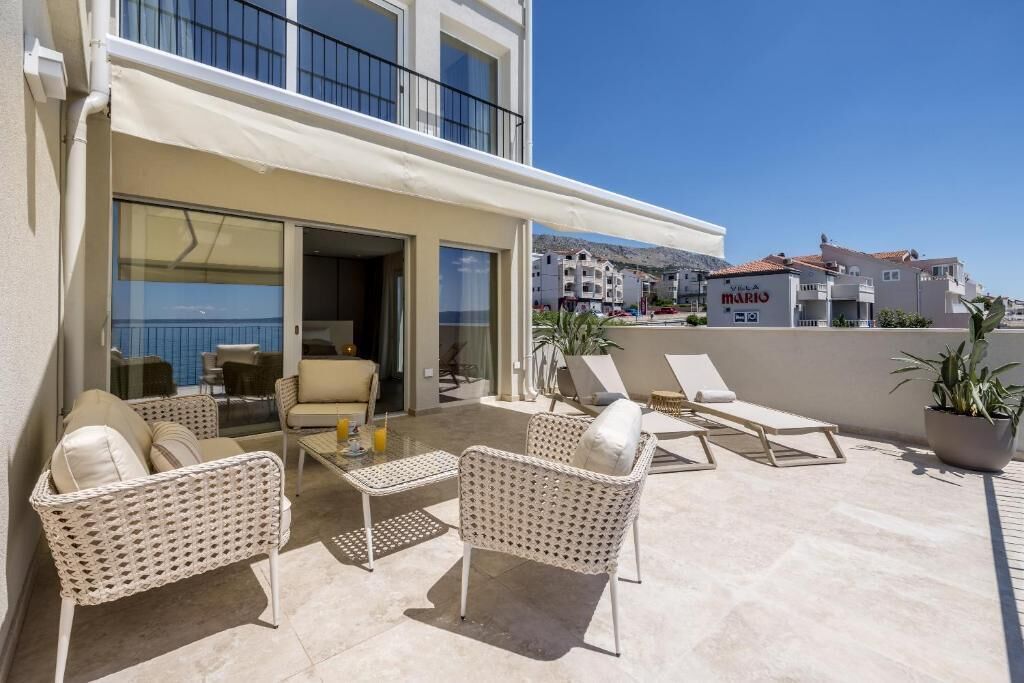 A balcony with wicker furniture, a glass door leading to an indoor area, and a view of the sea and buildings in the background.