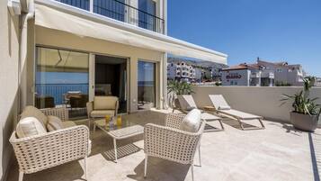 A balcony with wicker furniture, a glass door leading to an indoor area, and a view of the sea and buildings in the background.