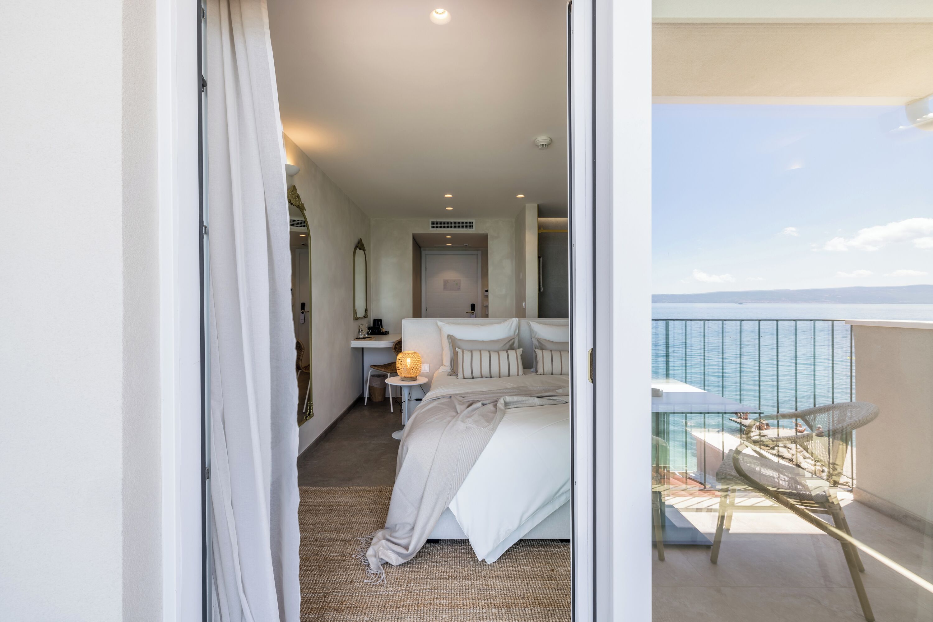 A bedroom with a view of the sea, a balcony with a table, and a small desk with a lamp.