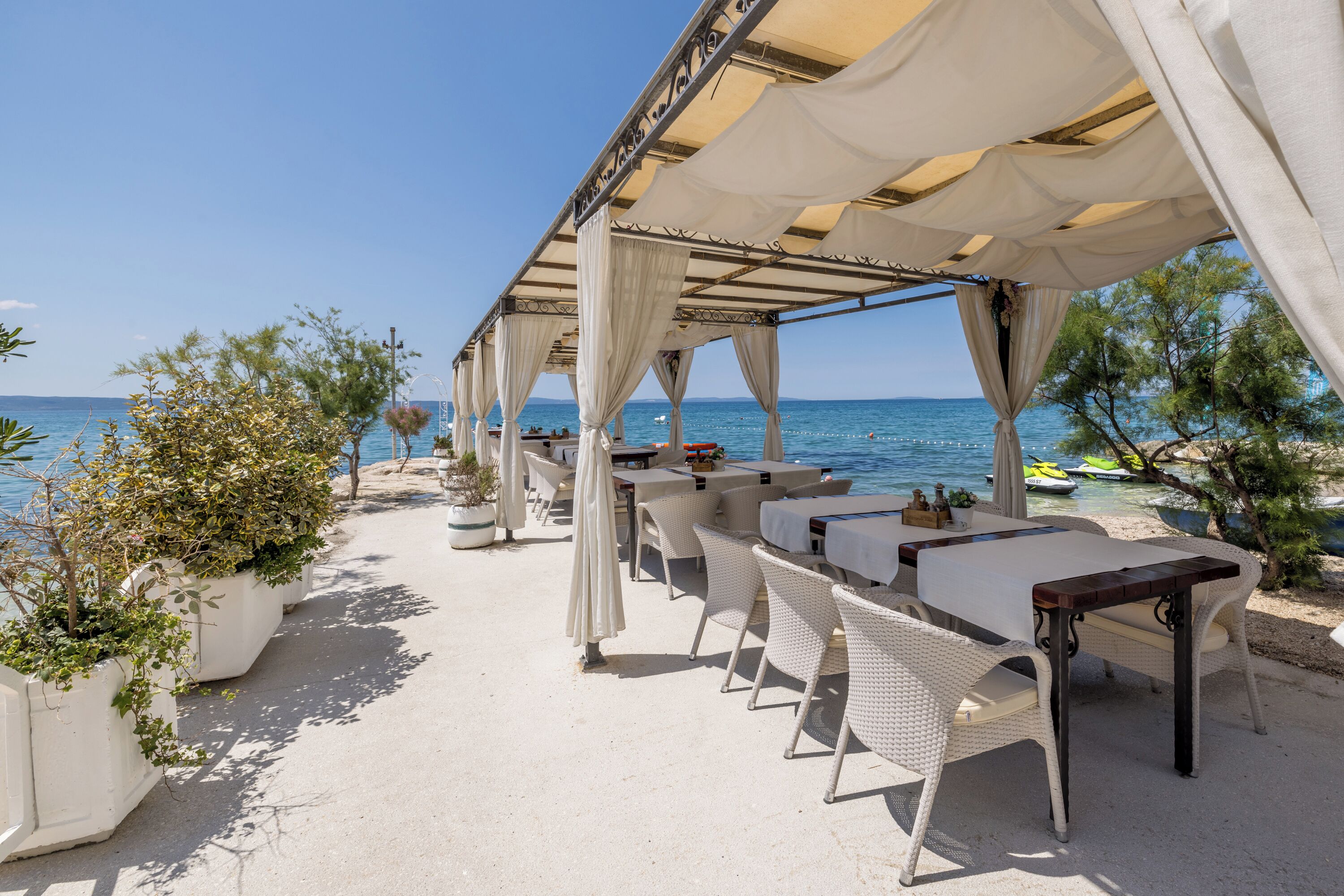 An outdoor seating area with tables and chairs, potted plants, and a view of the sea.
