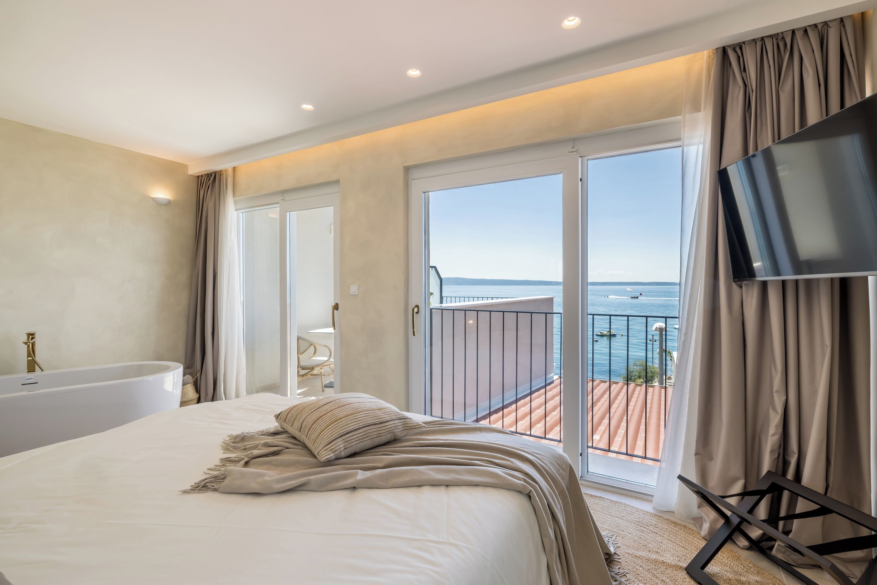A bedroom with a large bed, a TV, a bathtub, and a balcony with a view of the sea.