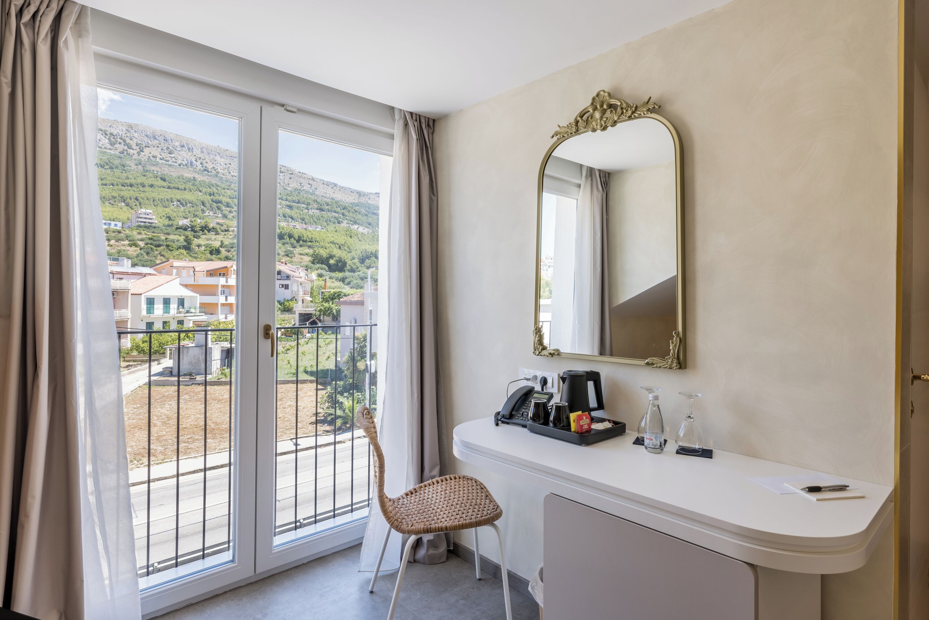 Ein Hotelzimmer mit Balkon, einem weißen Schreibtisch, einem Stuhl, einem Spiegel und Blick auf Gebäude und Hügel.