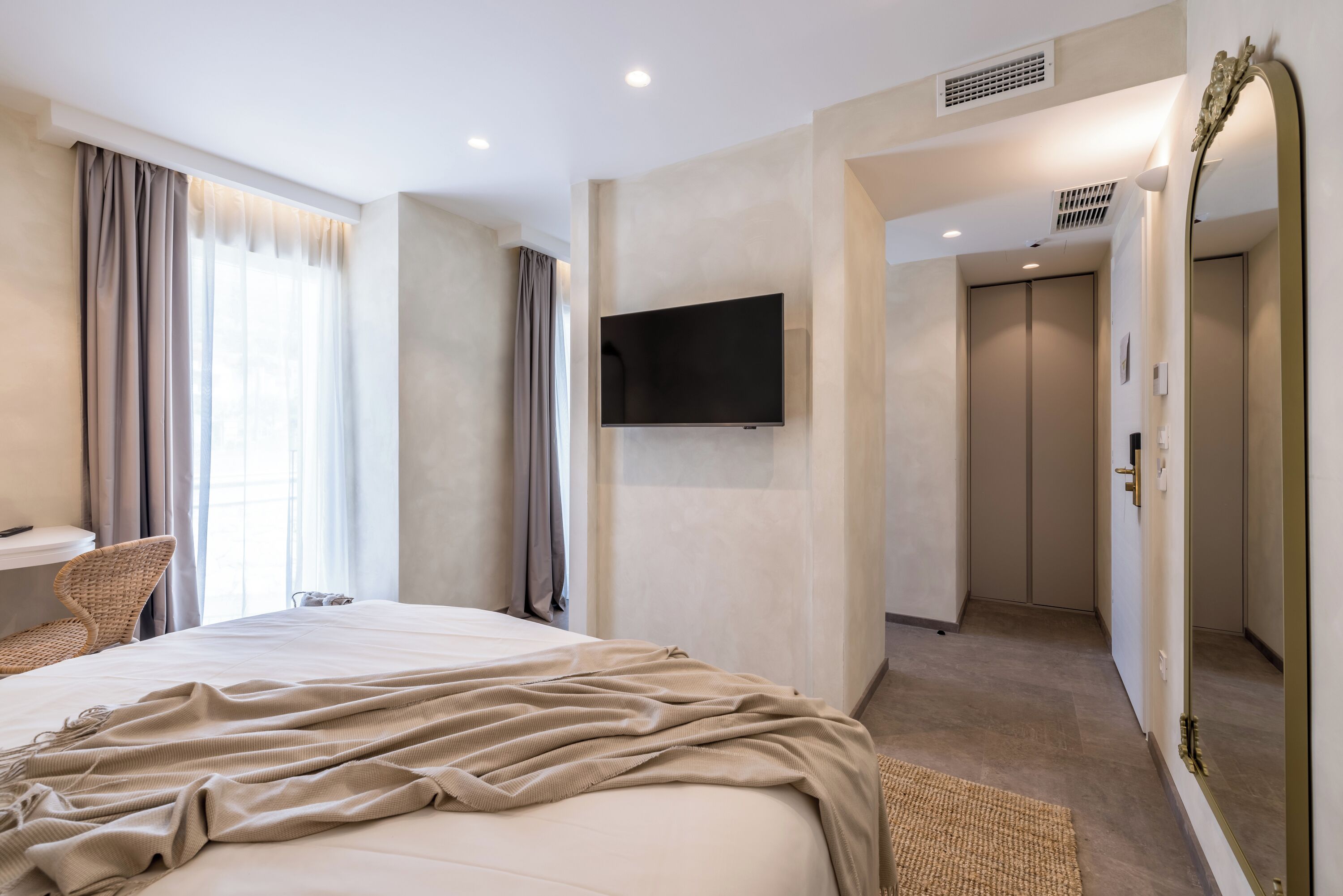 A modern hotel room with a large bed, a flat-screen TV mounted on the wall, a seating area with a chair, and a bathroom visible through an open door.