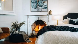 A bedroom with a fireplace, a bed with a white quilt, a dark throw blanket, a brown sofa, and a potted plant. - Oliver s Cottage Yarra Valley Spa and Sauna (Yarra Glen)