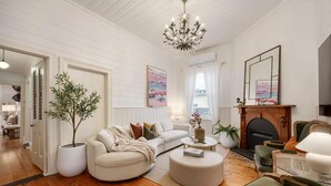 A living room with a white sofa, a round ottoman, a green armchair, a fireplace, and a chandelier. - Oliver s Cottage Yarra Valley Spa and Sauna (Yarra Glen)