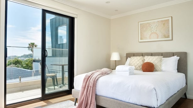A bedroom with a bed, a nightstand, a lamp, a window with a view of buildings and trees, and a sliding glass door to a balcony. - Balmain Family Tranquility - Harbour Views (Balmain East)