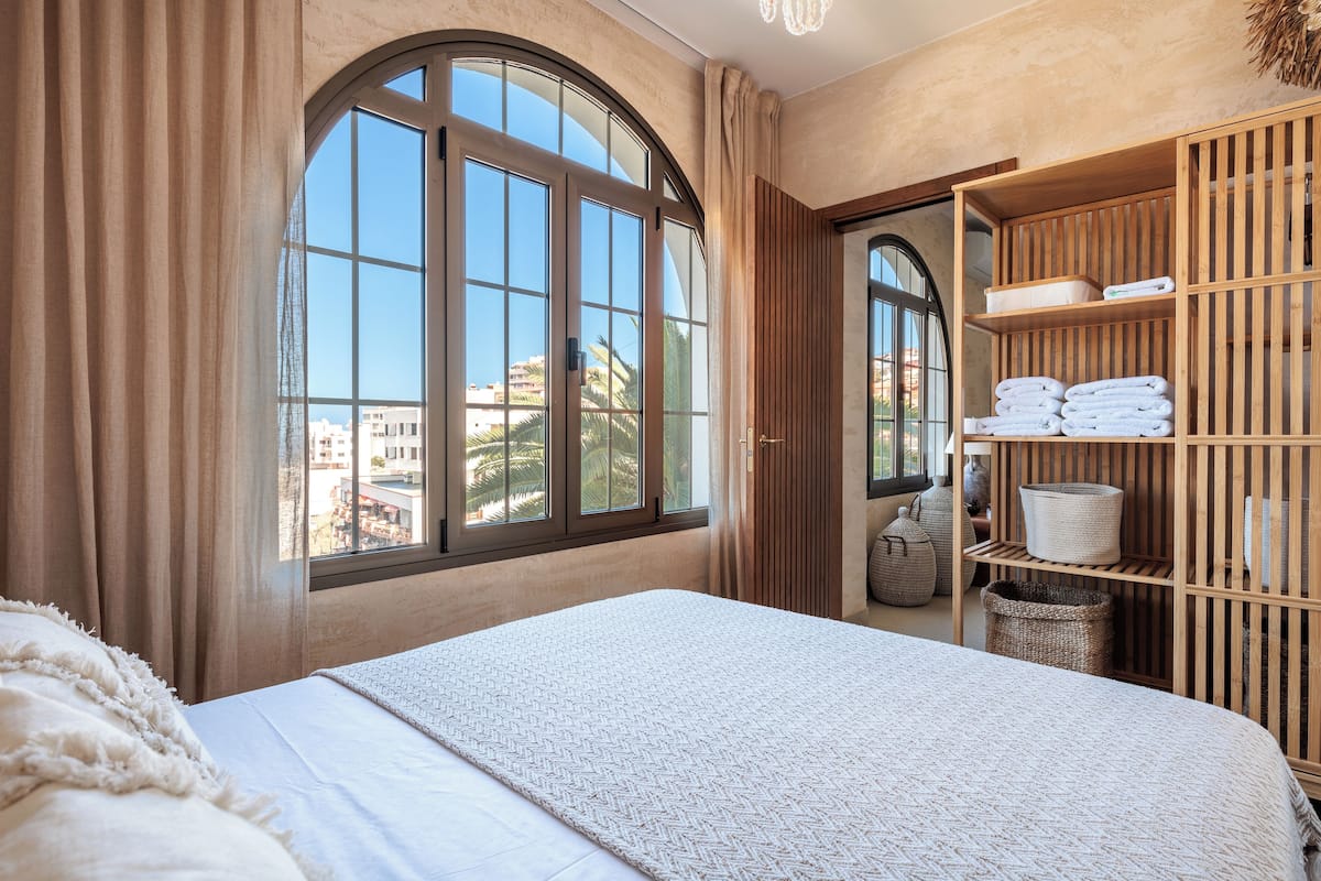 Un dormitorio con un amplio ventanal arqueado, una cama con colcha blanca y un estante de madera con toallas y cestas.