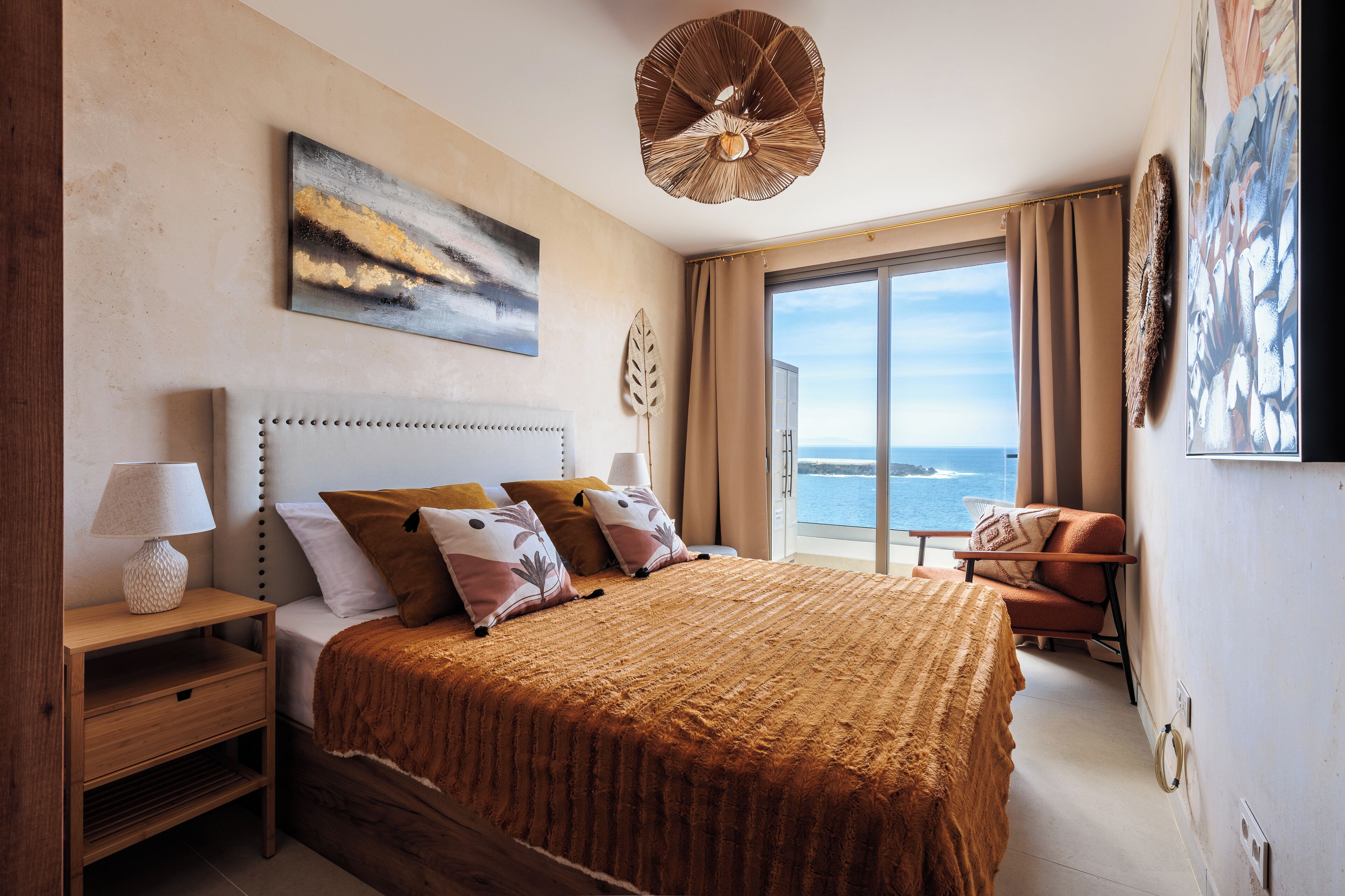 A bedroom with a large bed, a nightstand, a chair, and a view of the sea.