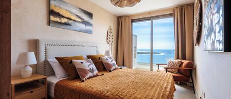 A bedroom with a large bed, a nightstand, a chair, and a view of the sea.