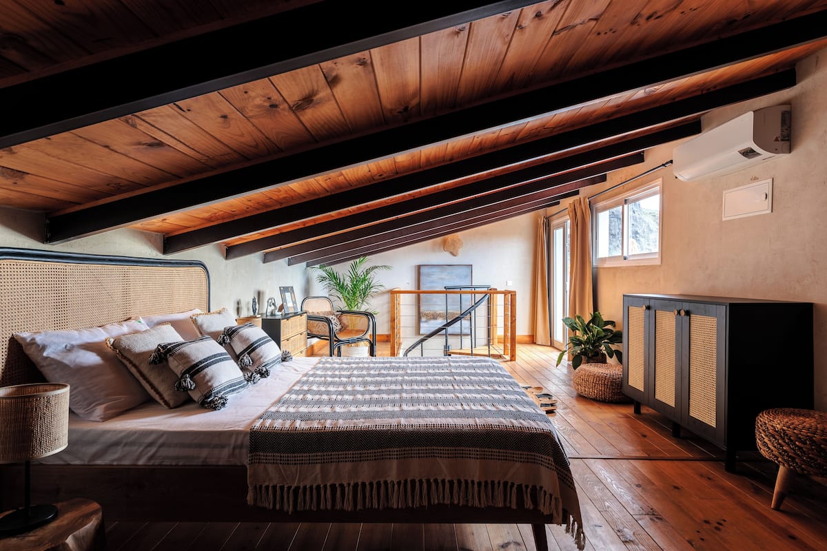 Un dormitorio con techo de madera, una cama con manta a rayas, una mesita de noche, una silla, una planta y vistas al exterior a través de una ventana.