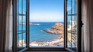 Vista costera a través de una ventana con cortinas, que muestra una costa rocosa, un edificio en un acantilado y un cielo despejado.