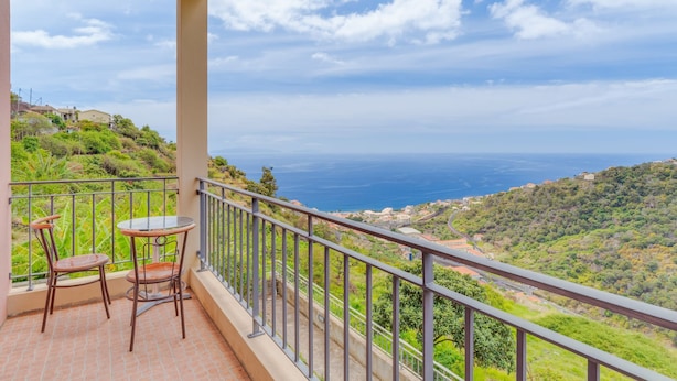 Un balcon avec deux chaises et une table, offrant une vue sur un paysage côtier avec des collines et la mer.