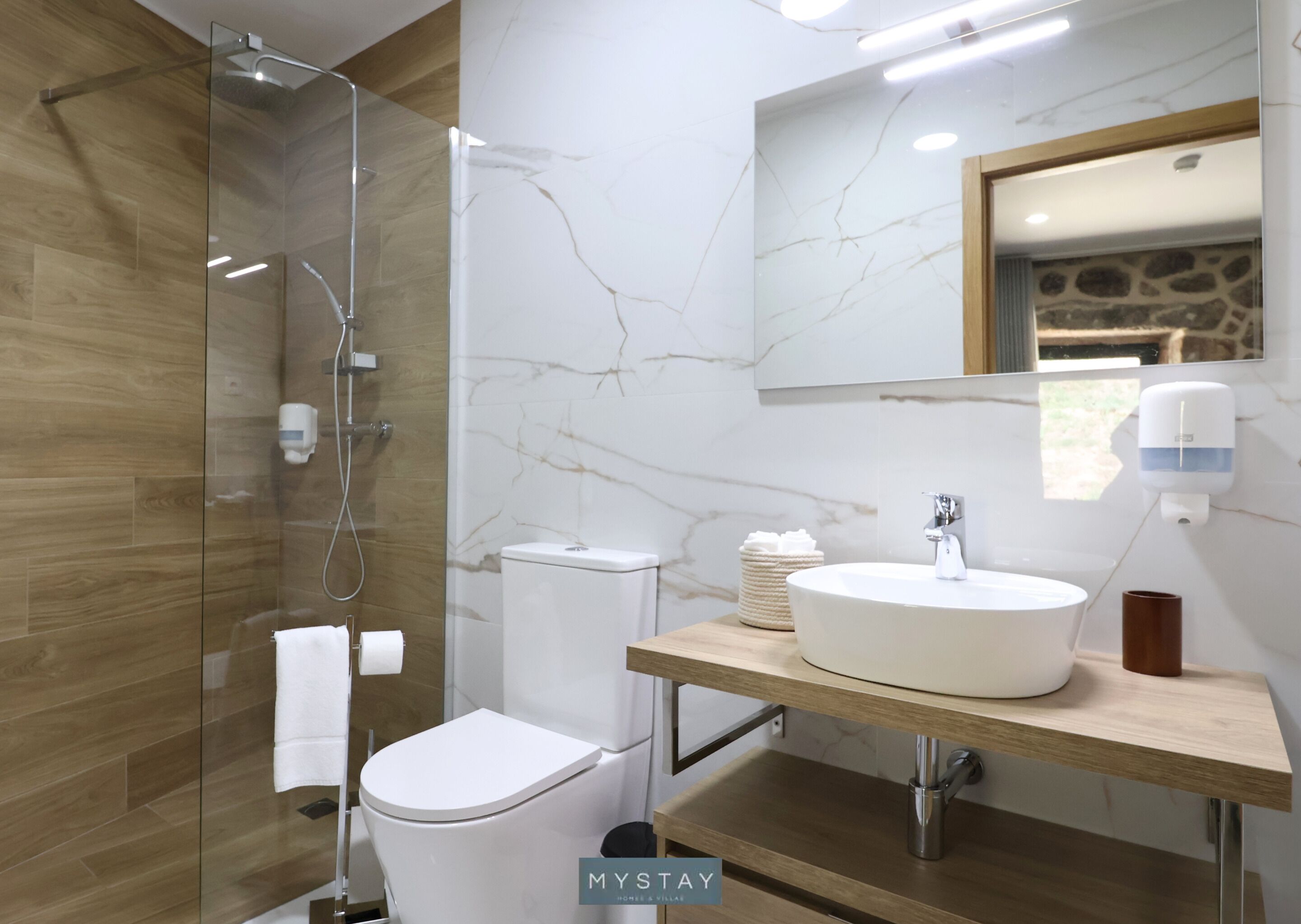 A modern bathroom with a glass shower enclosure, a white toilet, a marble-tiled wall, a wooden vanity with a sink, and a mirror above the vanity.