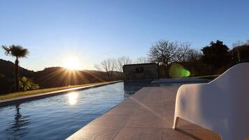 A swimming pool with a clear view of the sunset, a stone wall, and a palm tree.
