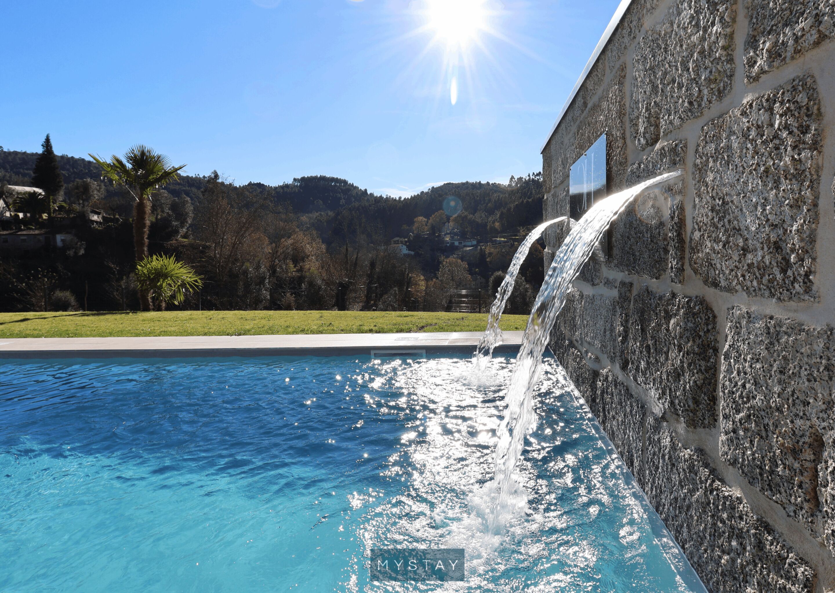 A swimming pool with a waterfall feature, surrounded by a stone wall and a grassy area with trees and hills in the background.