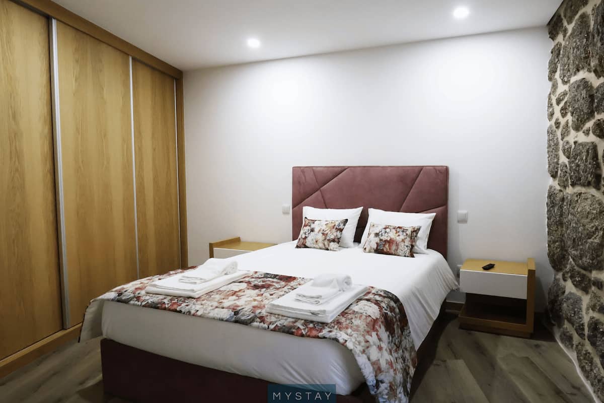 Ein ordentlich bezogenes Bett mit floralem Bettzeug, ein Holzschrank und eine Wand mit Steinverkleidung.