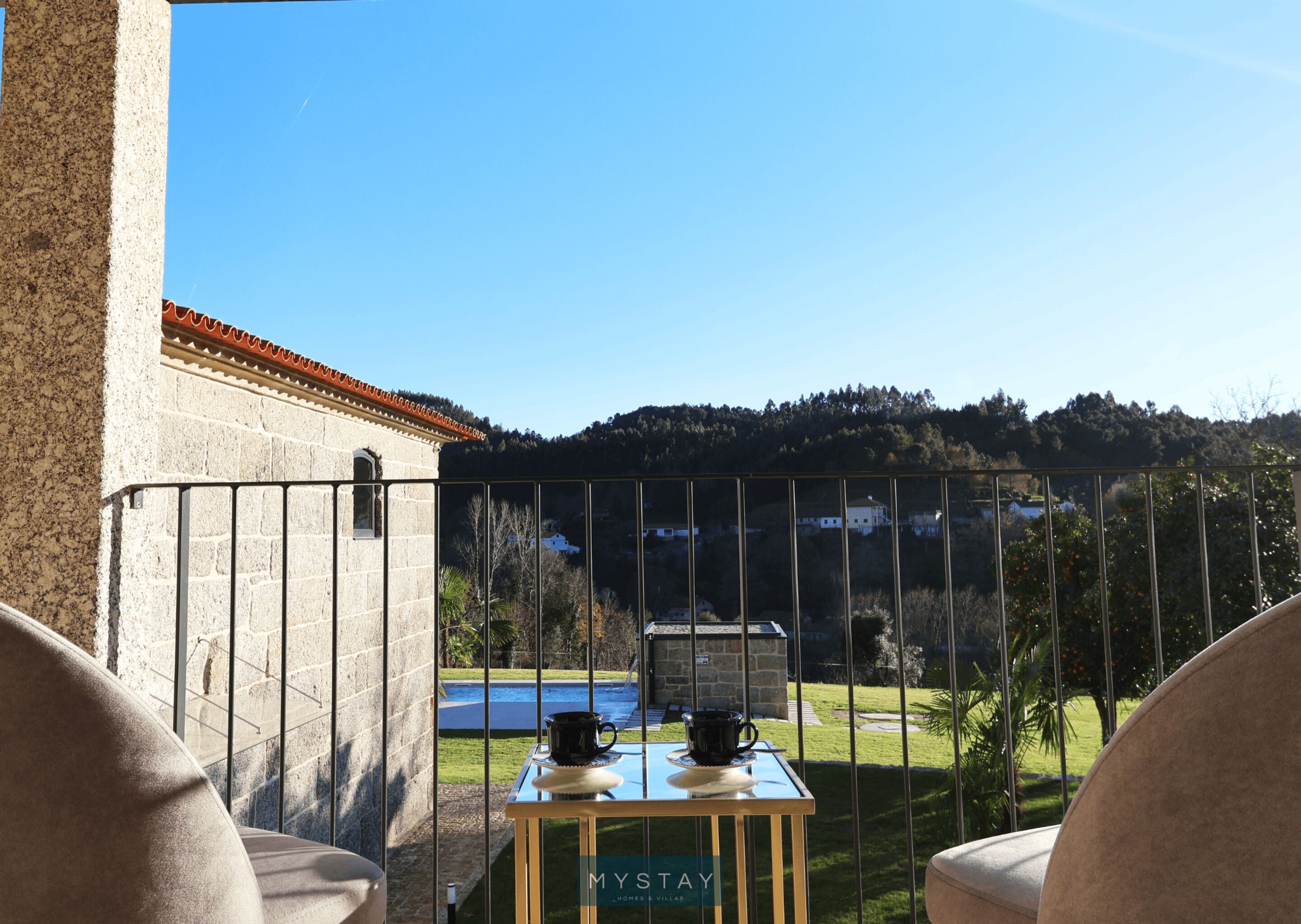 Ein Balkon mit Tisch, zwei Stühlen und Blick auf ein Schwimmbecken sowie Gebäude in der Ferne.