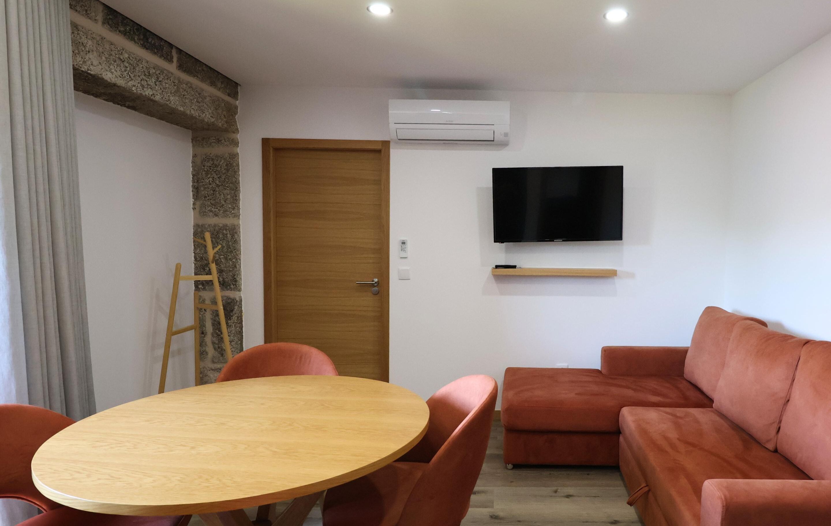 A room with a round wooden table, two orange chairs, a brown sofa, a television mounted on the wall, and a wooden door.