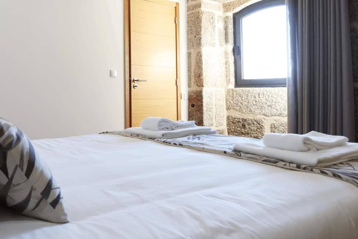 Ein ordentlich bezogenes Bett mit weißen Leinentüchern und einem gemusterten Kopfkissen, eine Holztür und eine Steinwand mit einem Fenster.