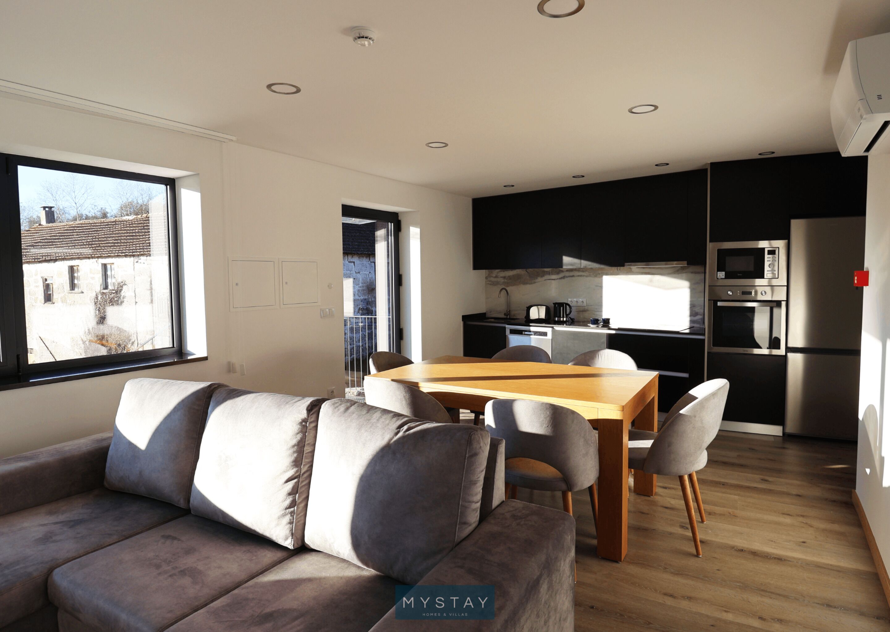 A modern living room with a sofa and a dining table, a kitchen with black cabinets and stainless steel appliances, and a large window with a view of buildings.