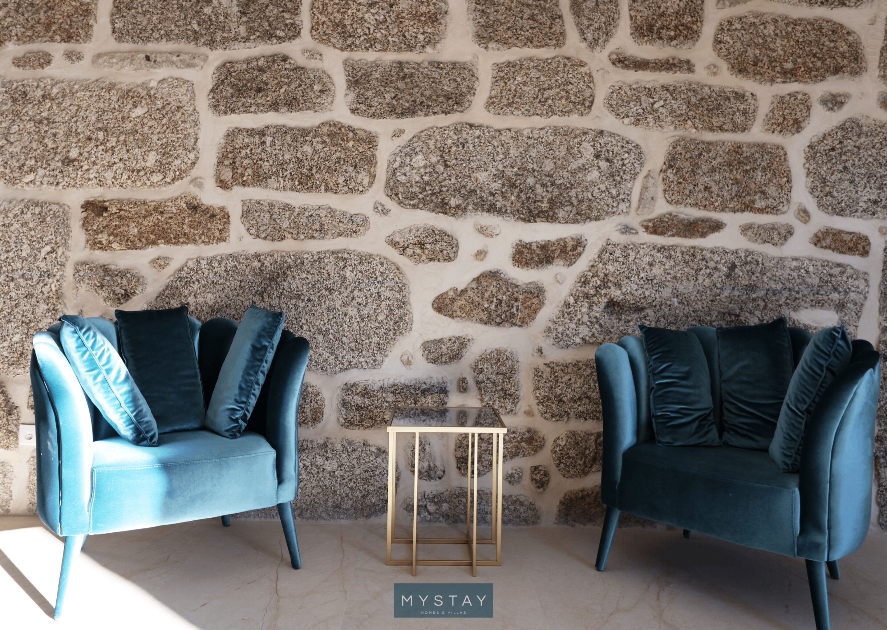 Two blue armchairs with cushions against a stone wall.