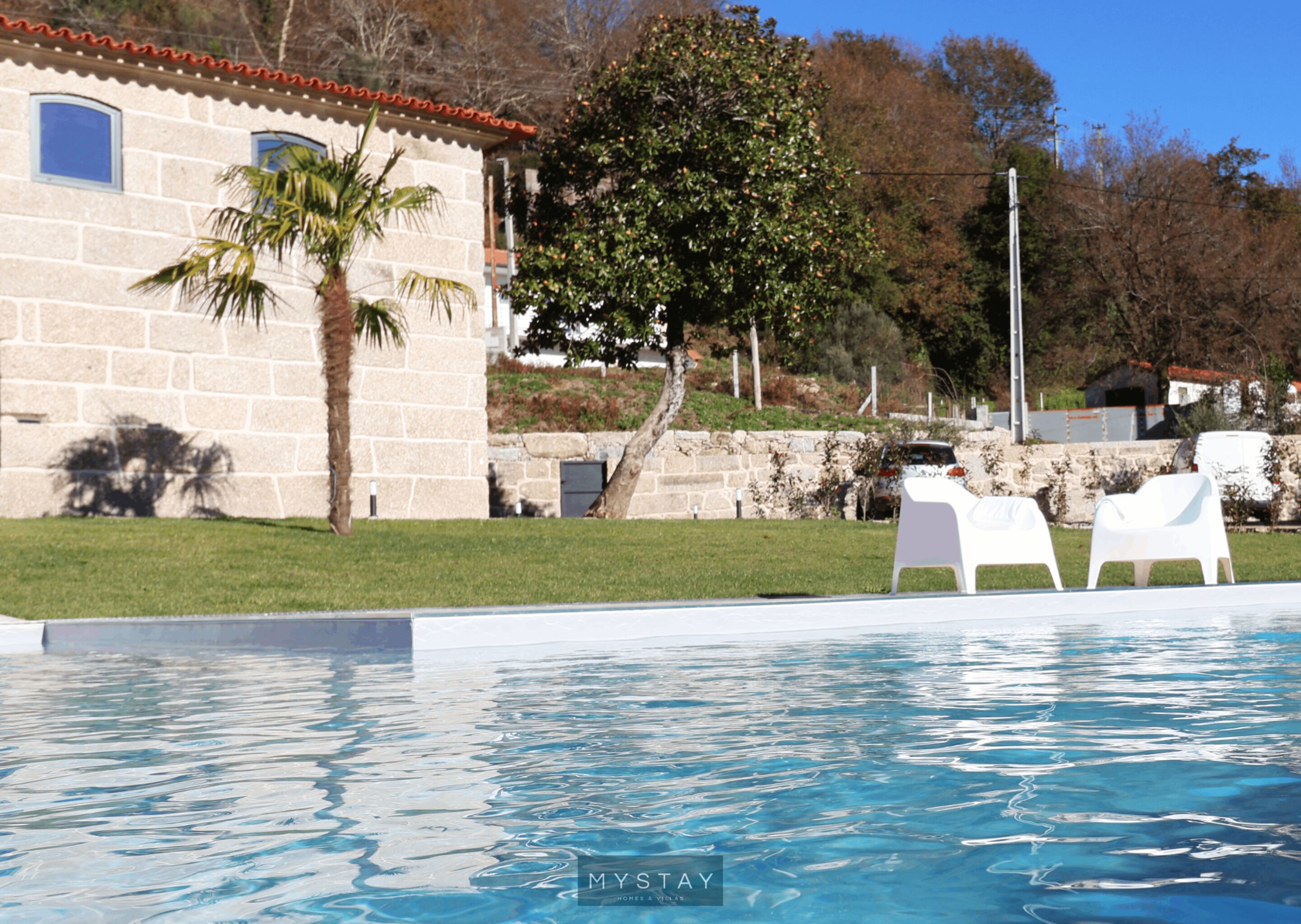 A swimming pool with white lounge chairs, a stone building with windows, and a palm tree.