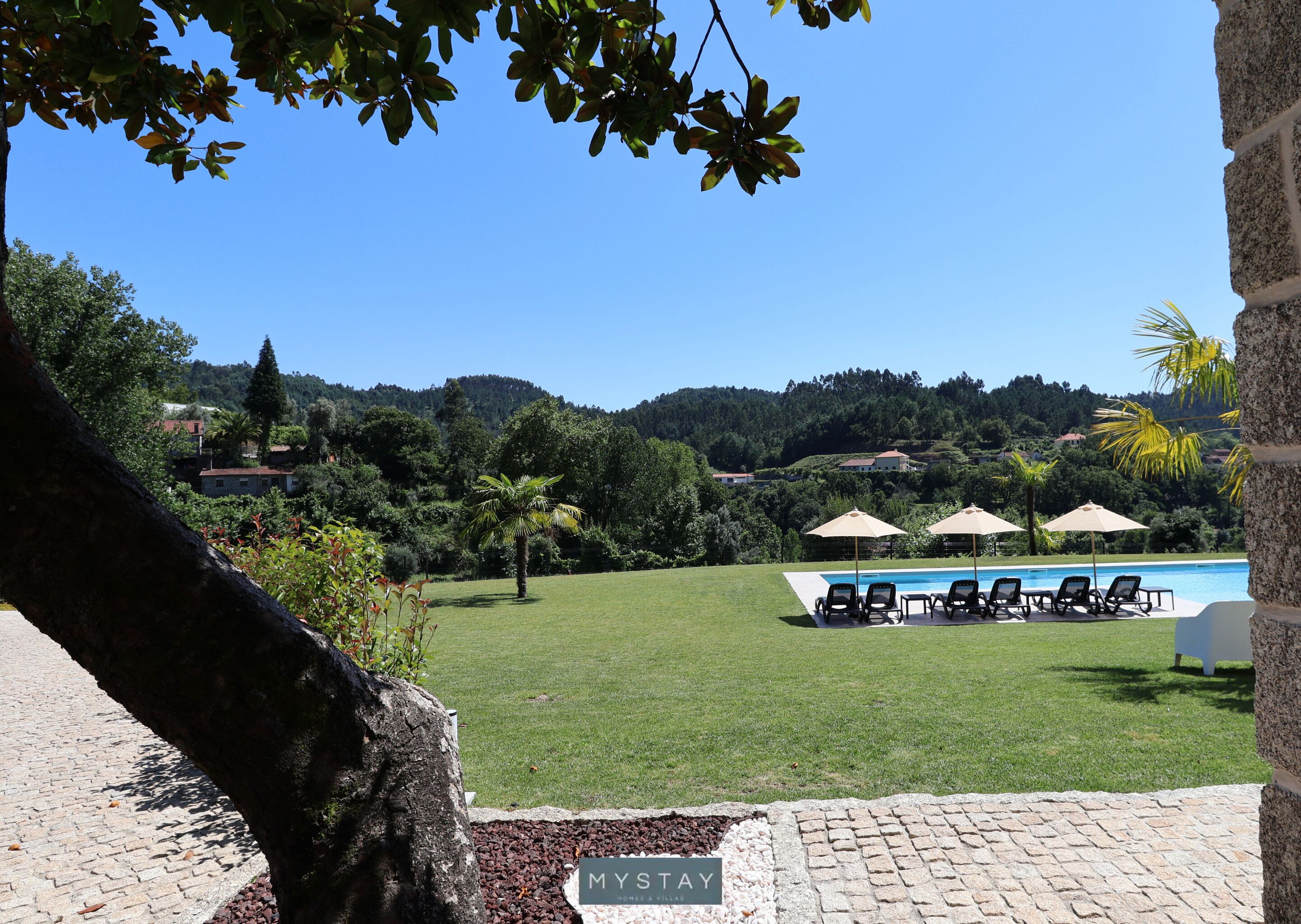 A pool area with lounge chairs and umbrellas, surrounded by greenery and a view of hills.