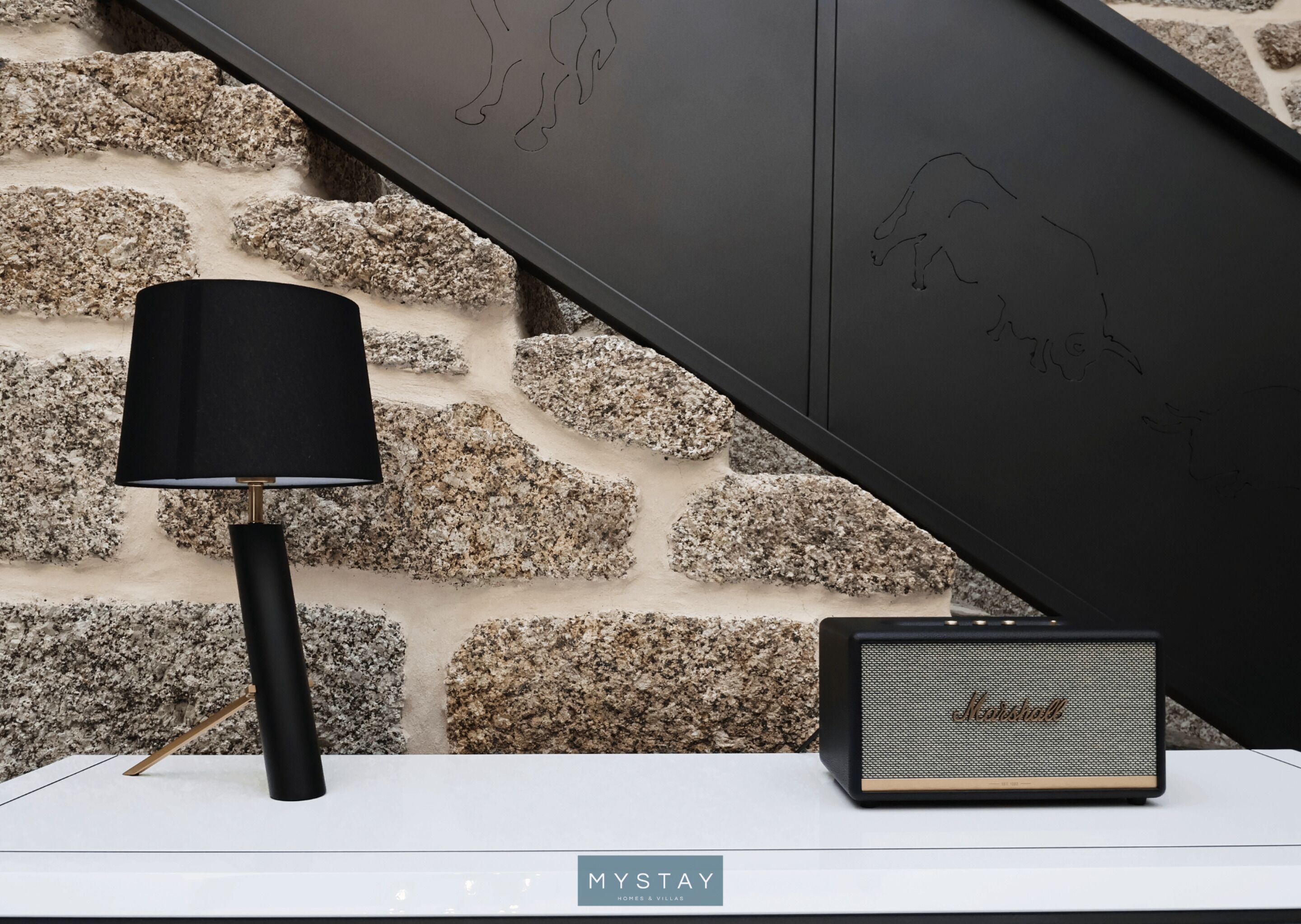 Une lampe et un haut-parleur sur une surface blanche, devant un mur de pierre orné de dessins abstraits d’animaux.