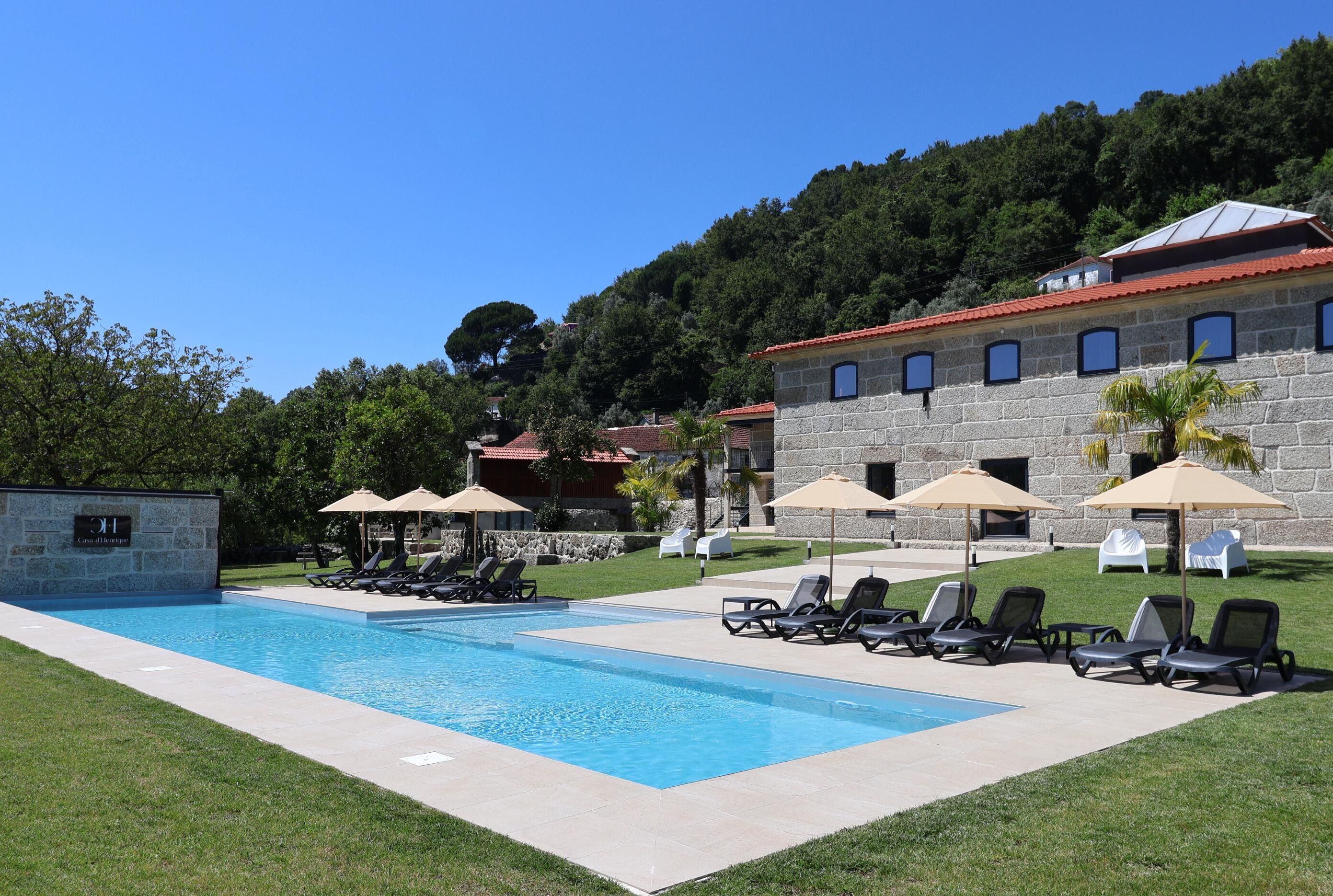 A swimming pool with lounge chairs and umbrellas, surrounded by a stone building with a red roof, set against a hillside.
