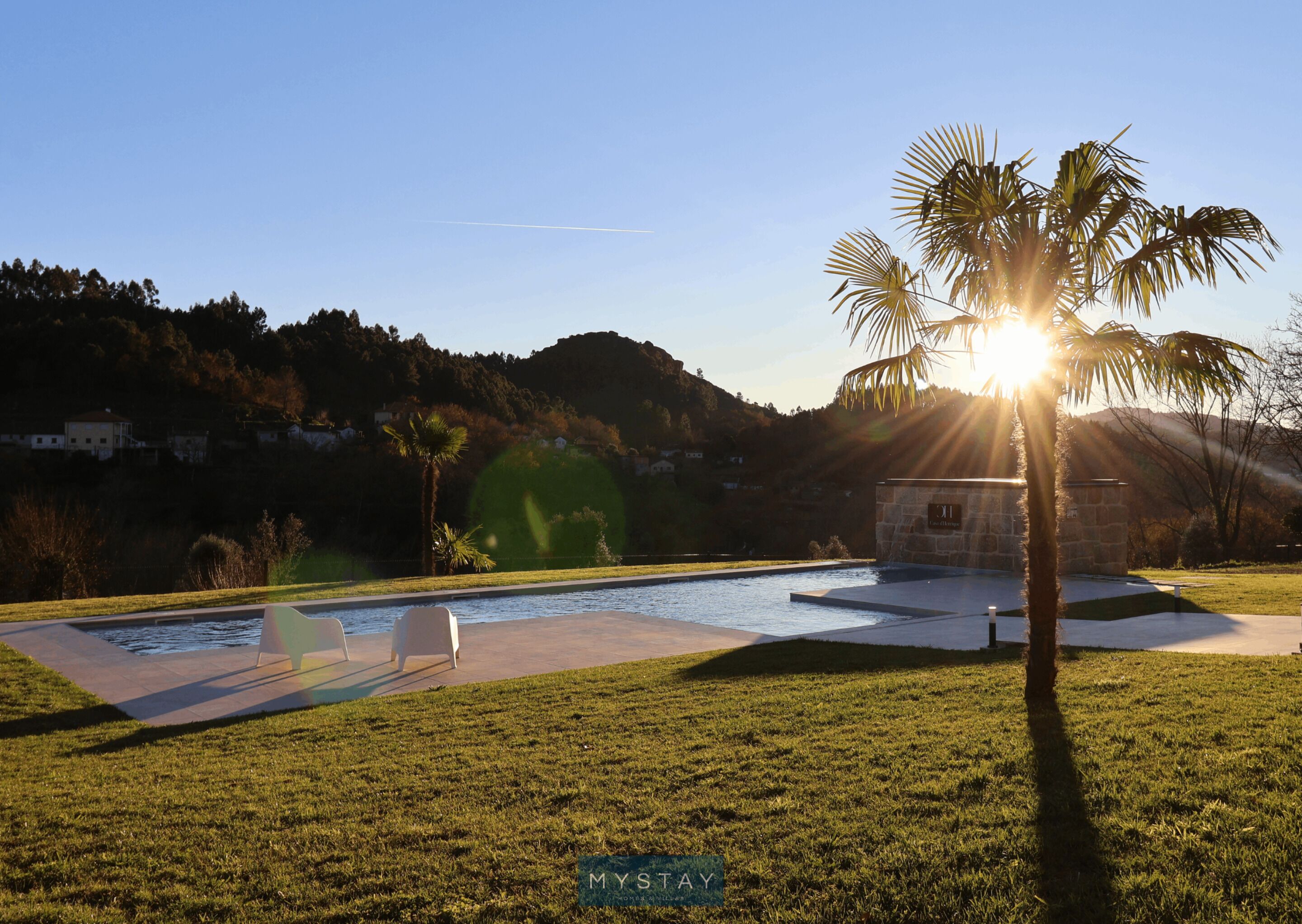 A swimming pool with a stone structure labeled "MYSTAY" in the background, surrounded by grass and palm trees.