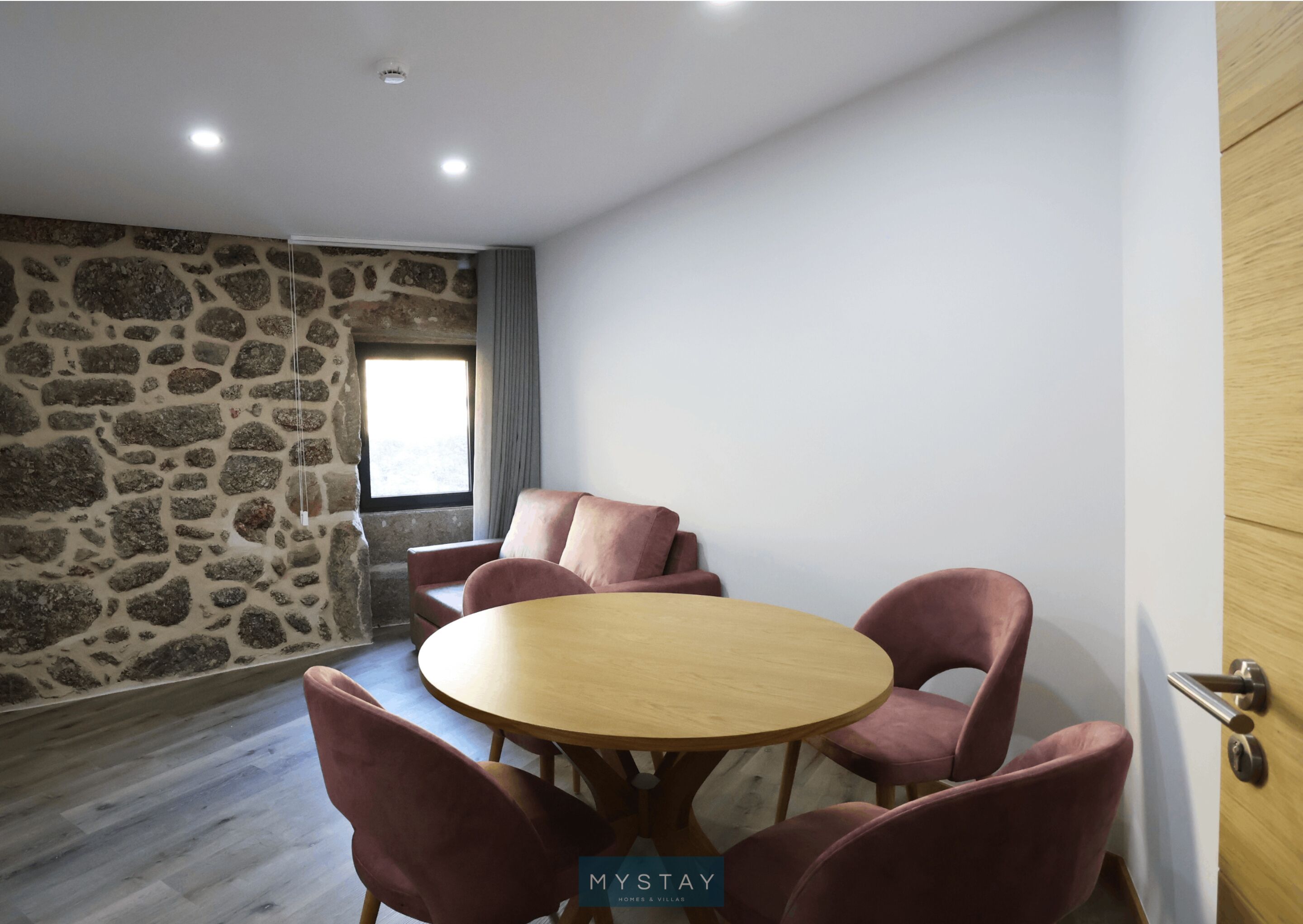 A room with a stone wall, a round wooden table with six chairs, a window with curtains, and a door with a handle.