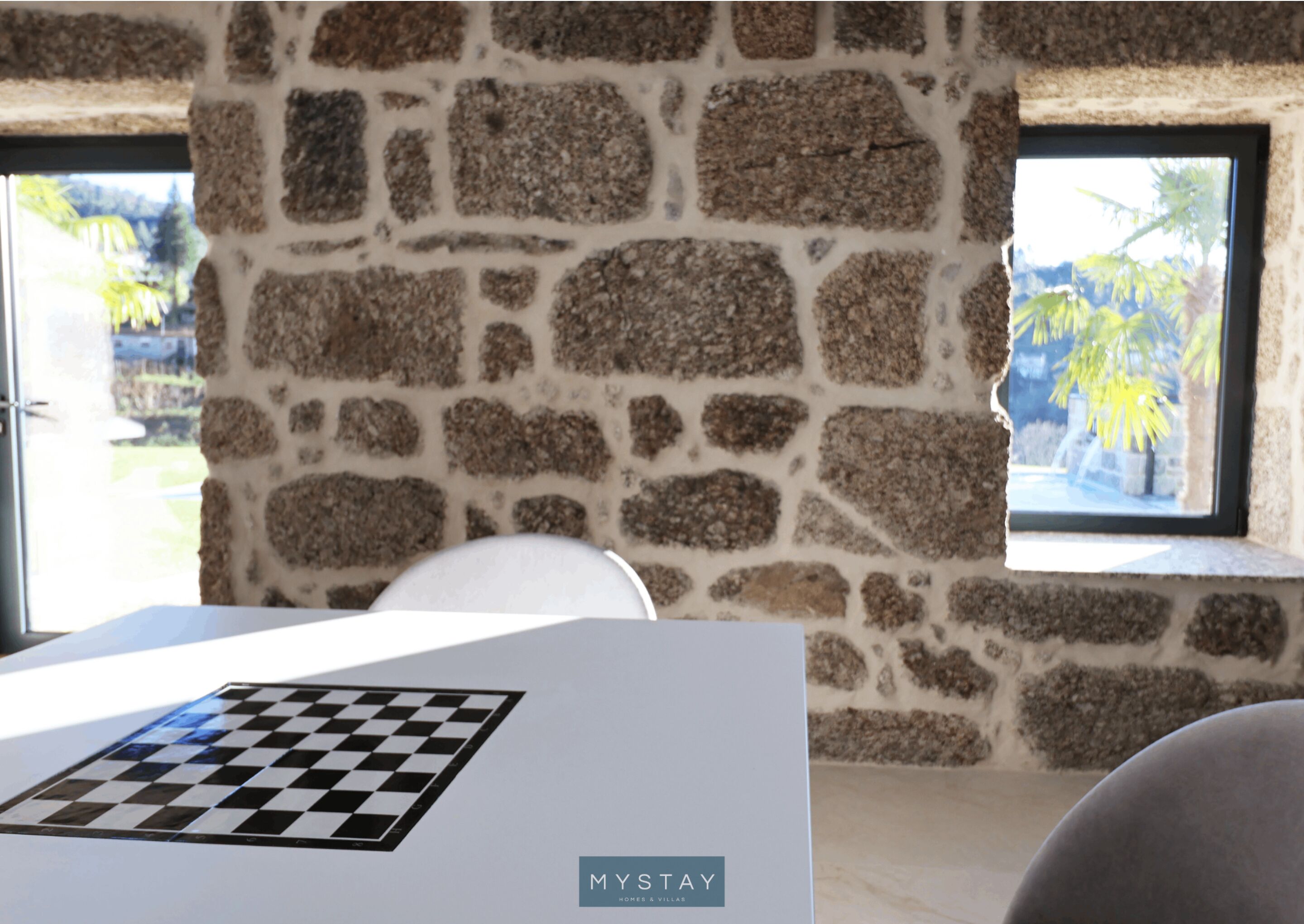 A room with a stone wall, a window with a view of palm trees, and a chessboard on a table.