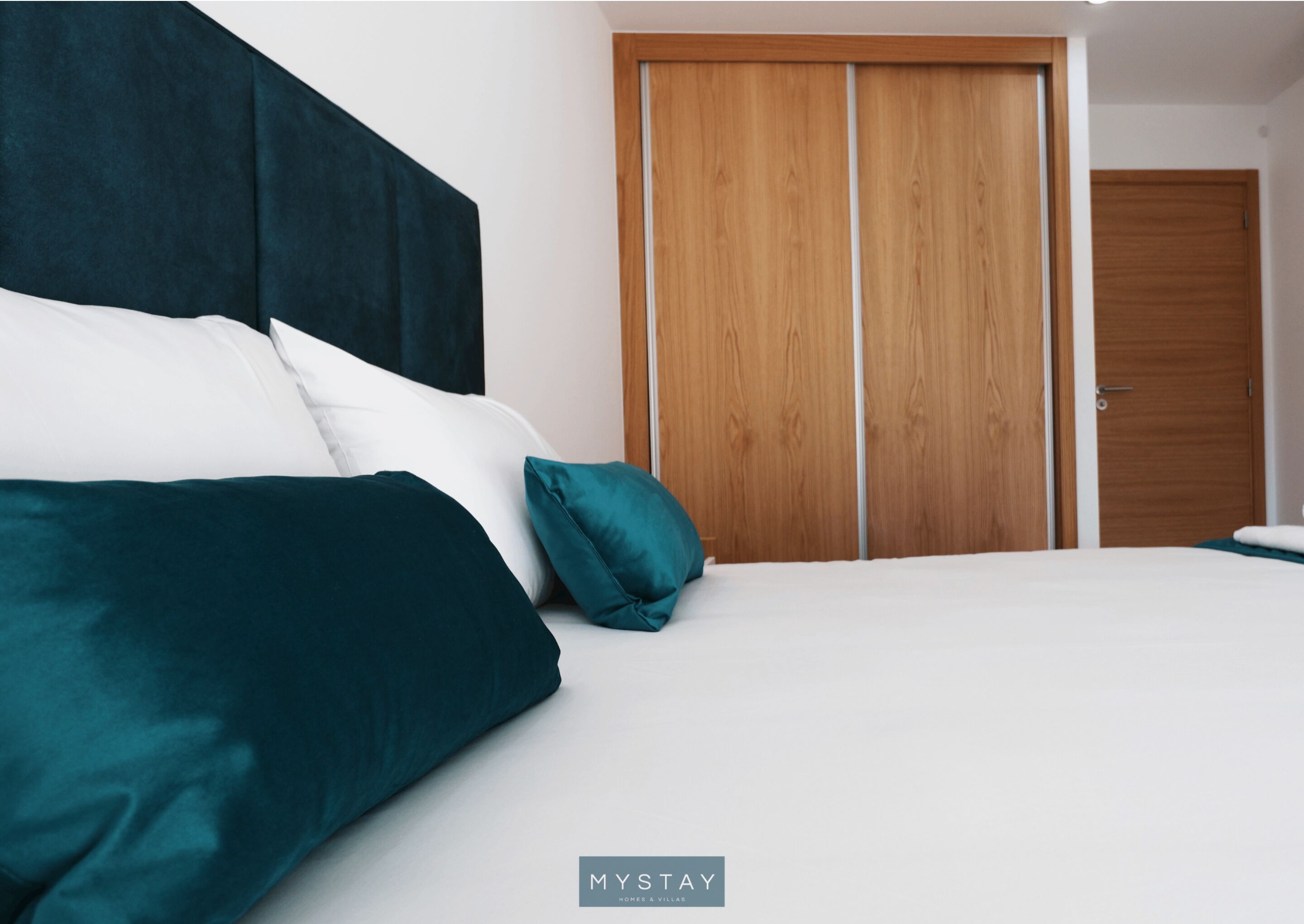 A bedroom with a bed, dark headboard, white bedding, teal pillows, and a wooden wardrobe.