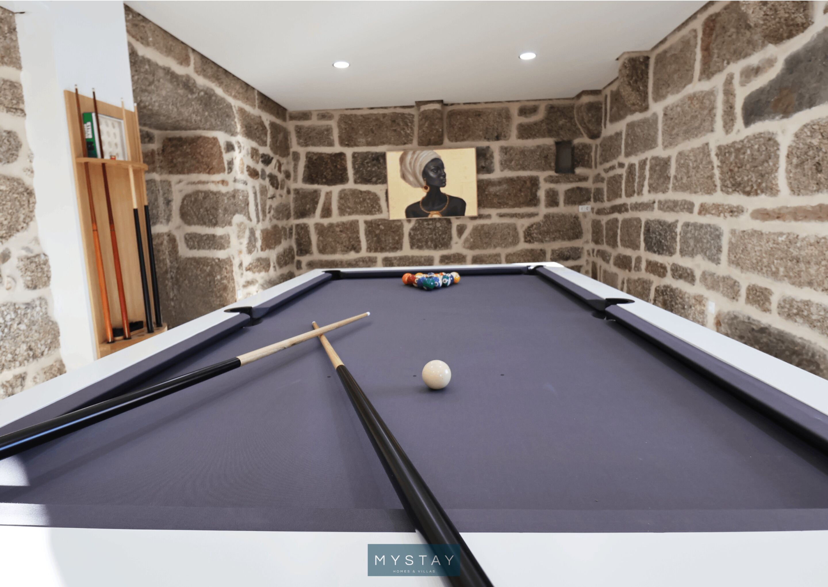 Une table de billard avec une boule blanche et des queues de billard, placée devant un mur de pierre orné d’un tableau encadré.