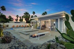 Un espace de vie extérieur moderne comprenant une piscine, des chaises longues, un foyer et un espace repas fermé par des parois de verre.