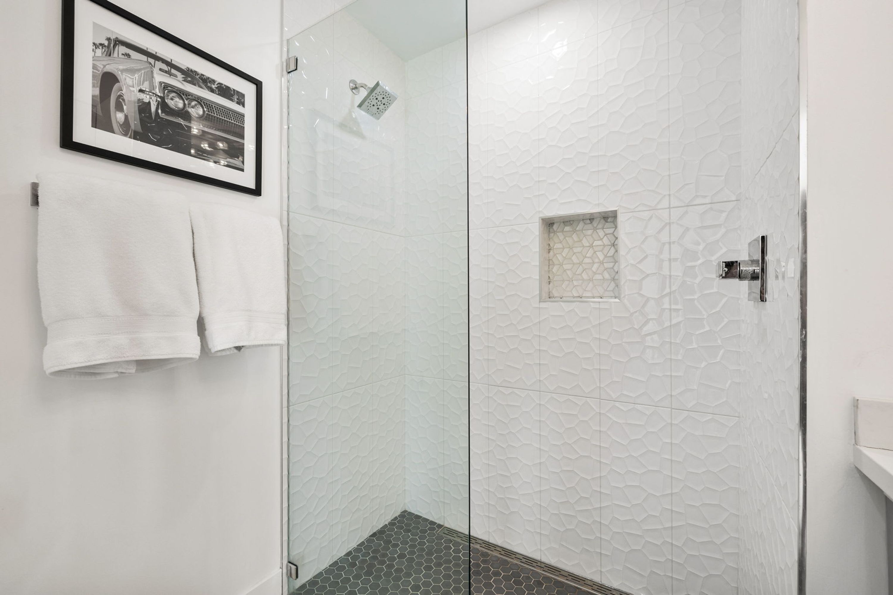 Une salle de bain moderne avec une douche à parois de verre, des murs blancs à texture et une photographie encadrée accrochée au mur.