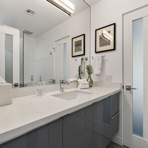 Un bagno moderno con uno specchio grande, un lavandino bianco e mobili scuri. Due opere d'arte incorniciate sono appese alla parete e un vaso con pianta si trova sul ripiano.