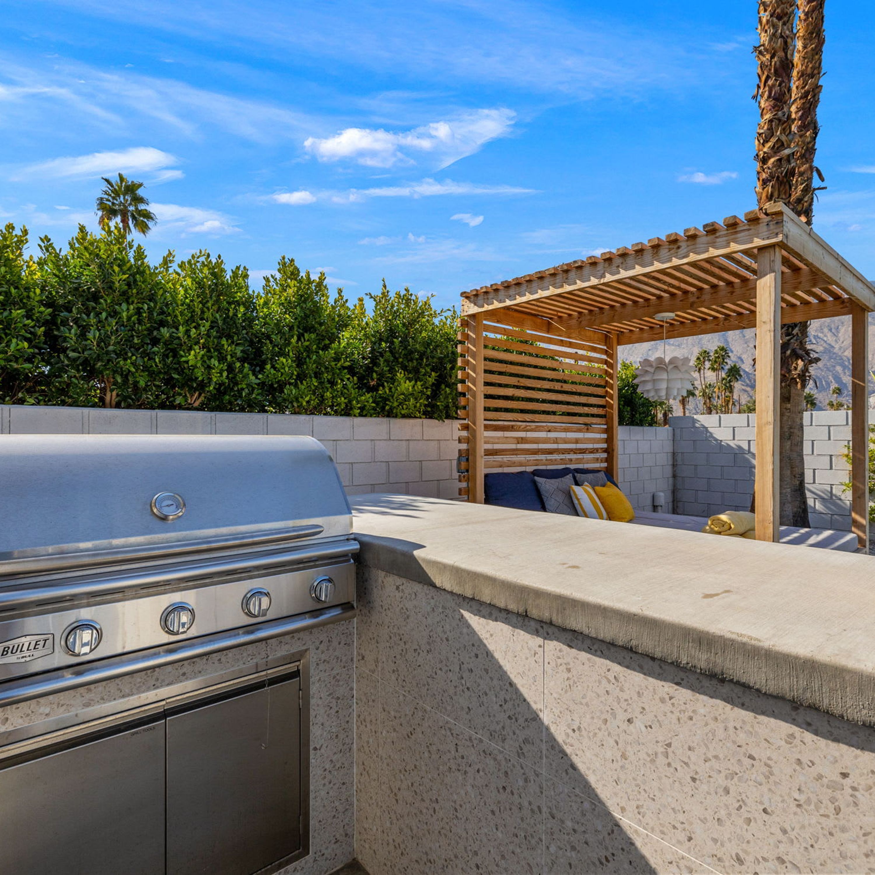 An outdoor kitchen with a built-in grill, a wooden pergola, and a seating area with cushions.