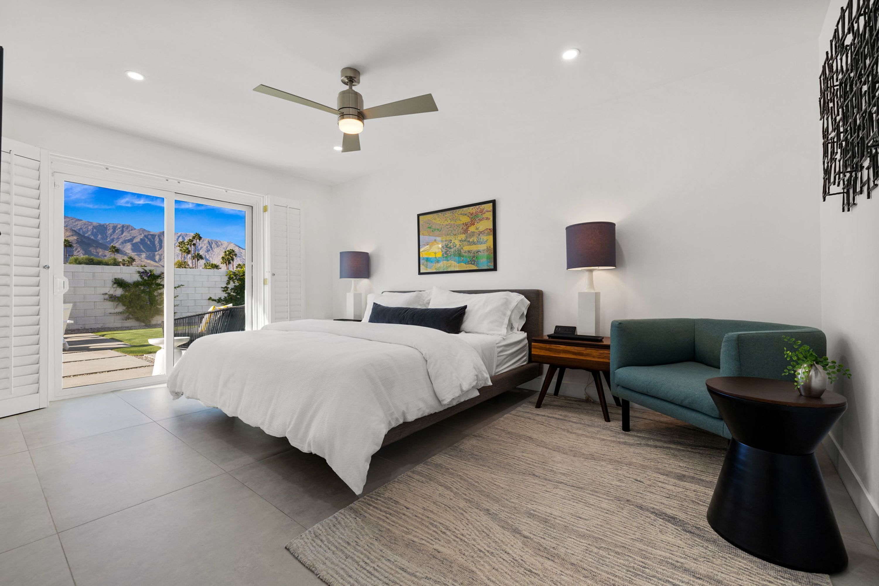 A bedroom with a bed, a green armchair, a side table, a ceiling fan, and a painting on the wall.